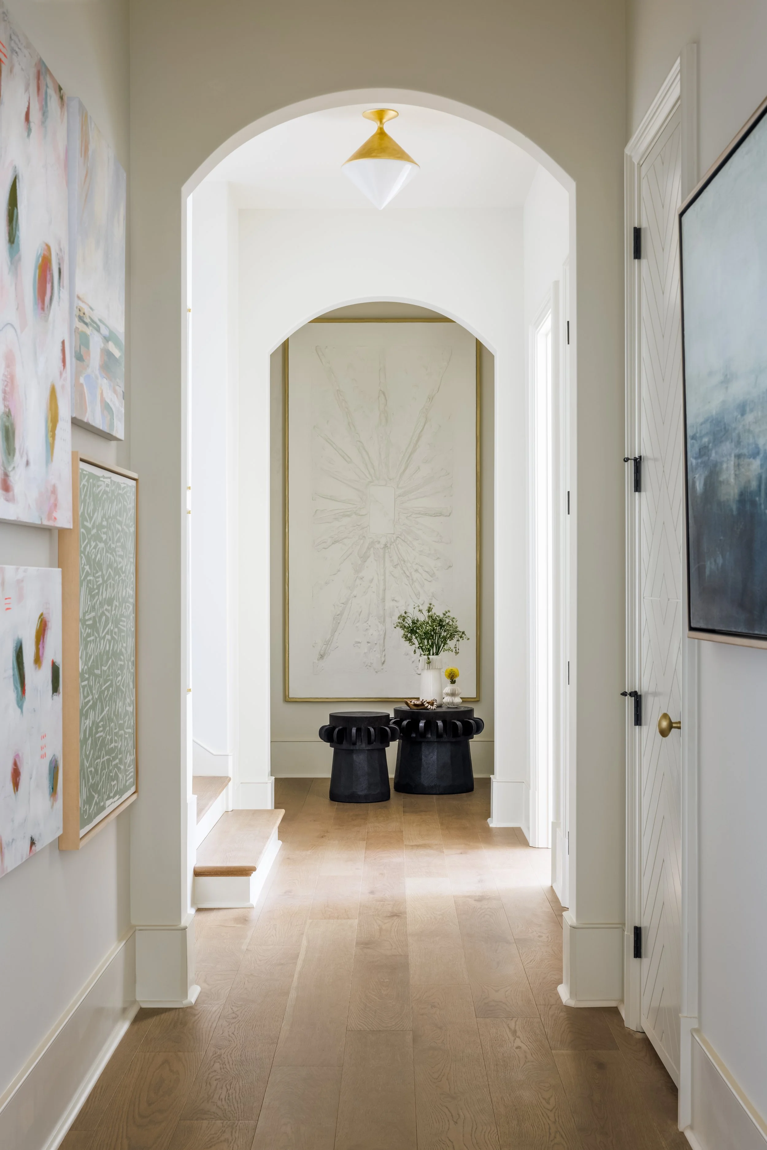 Bright arched hallway with light wood floors, colorful artwork, a statement piece at the end of the corridor, and black sculptural stools.