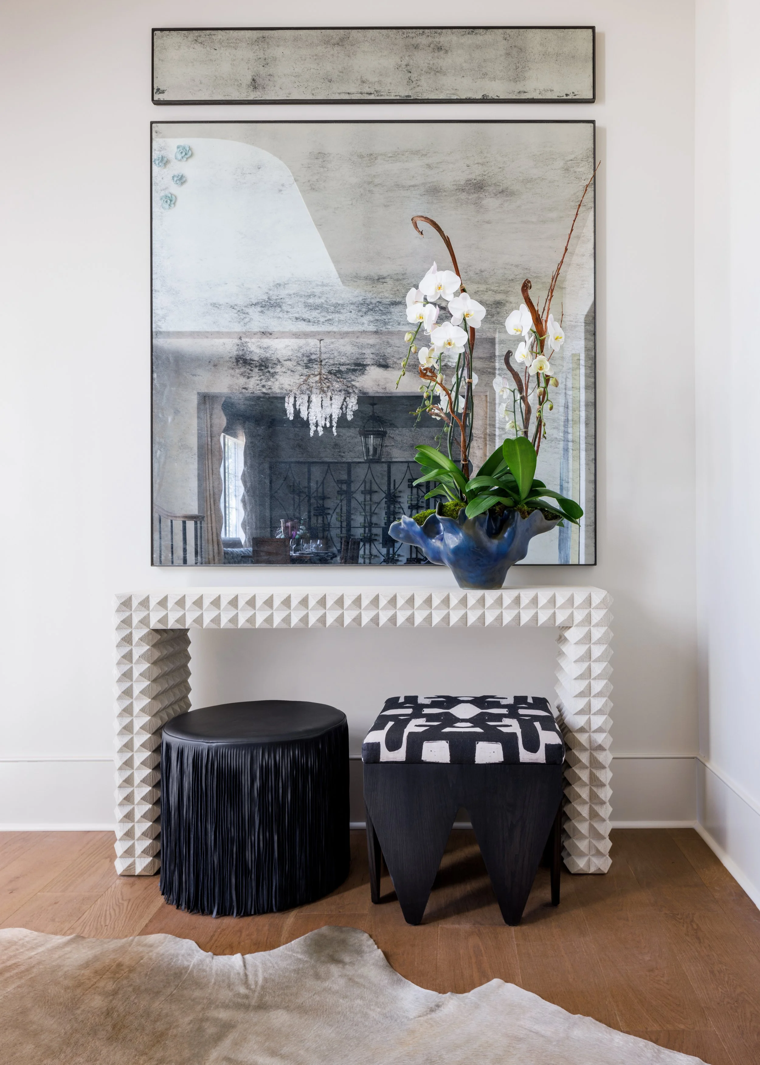 Decorative console vignette with a large abstract mirror, white orchids, sculptural stools, and layered black-and-white accents.