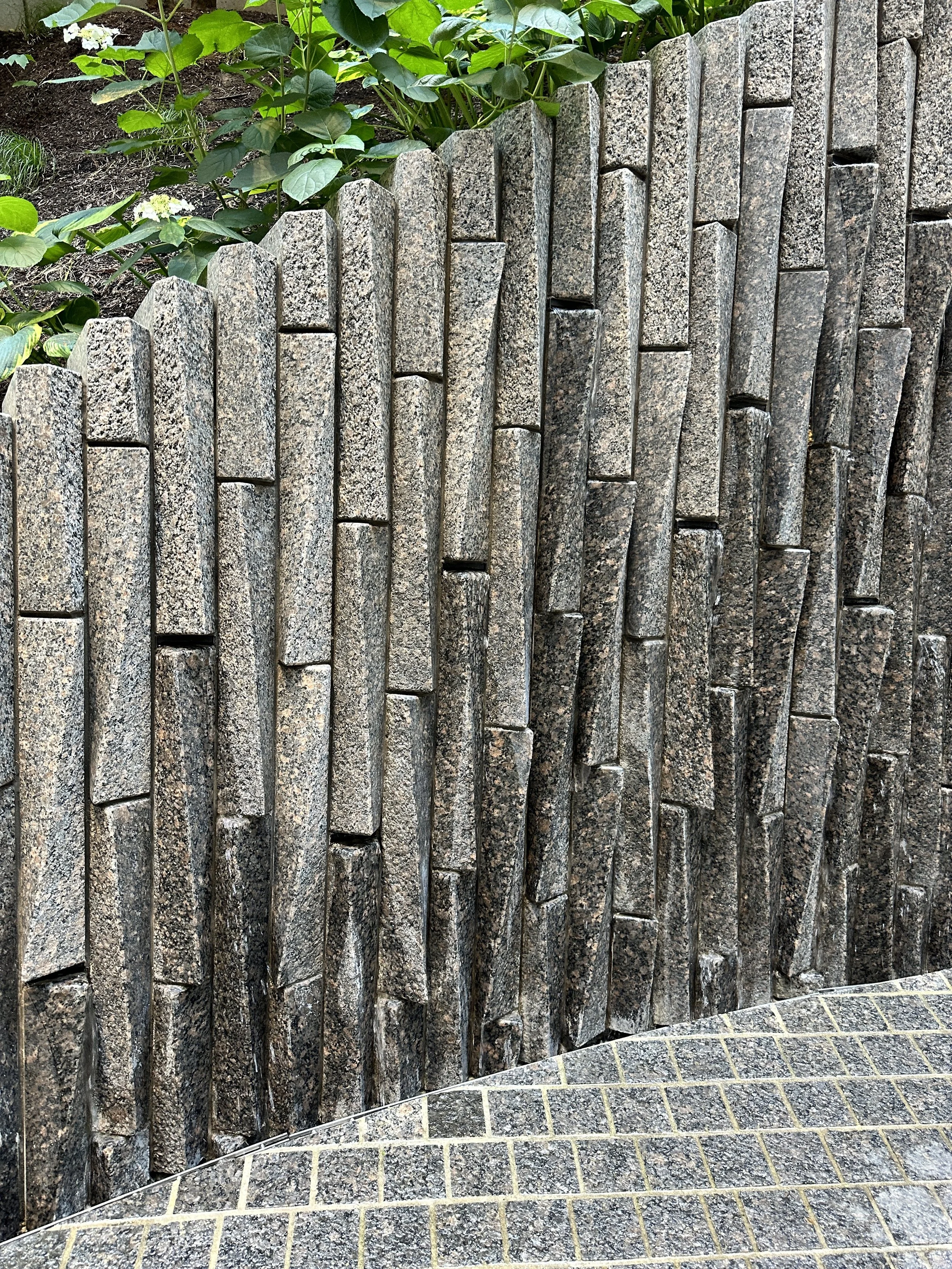 Textured vertical stone wall beside patterned paving and greenery.