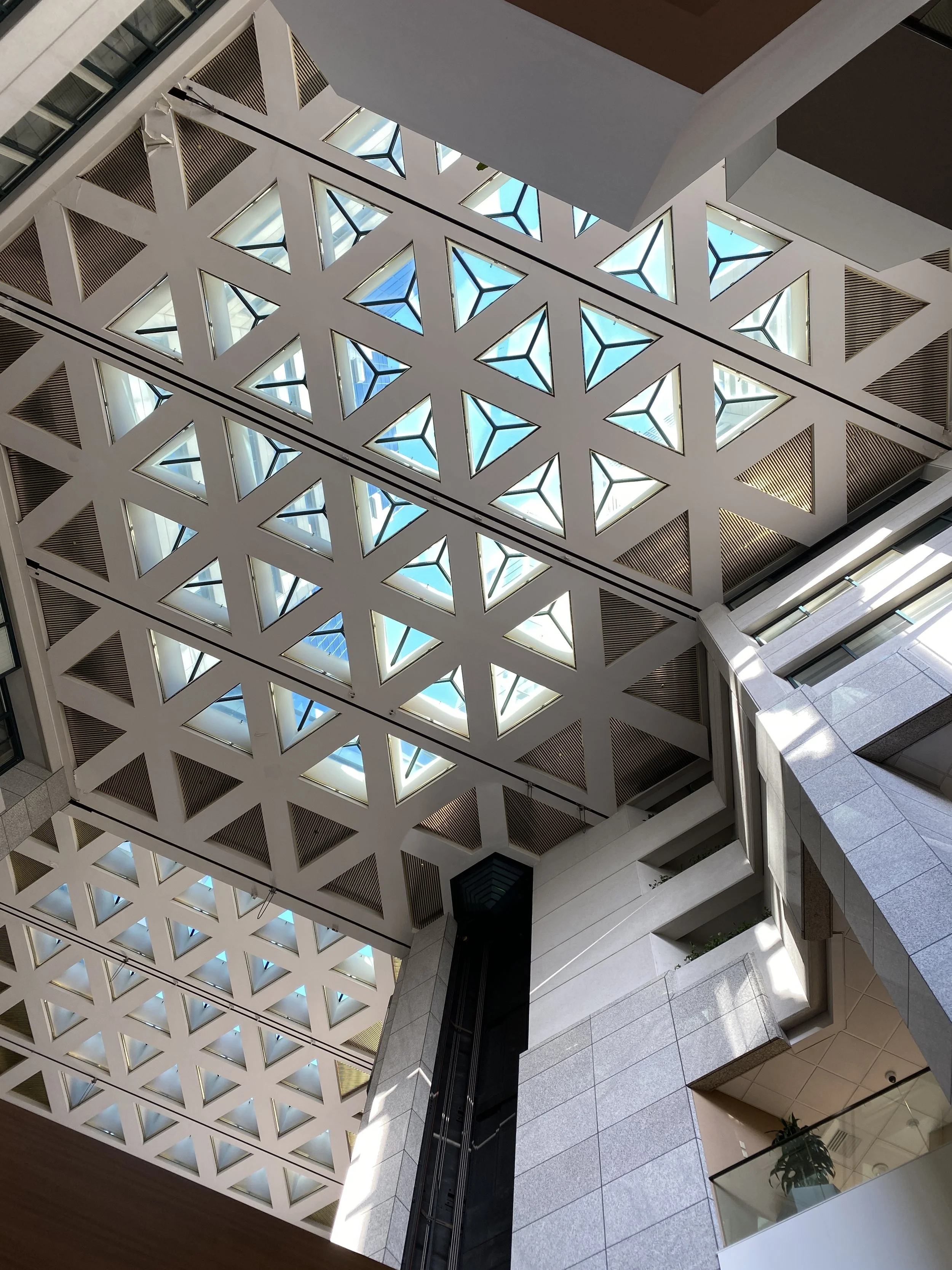 Geometric architectural ceiling with repeating triangular openings and blue sky visible above.