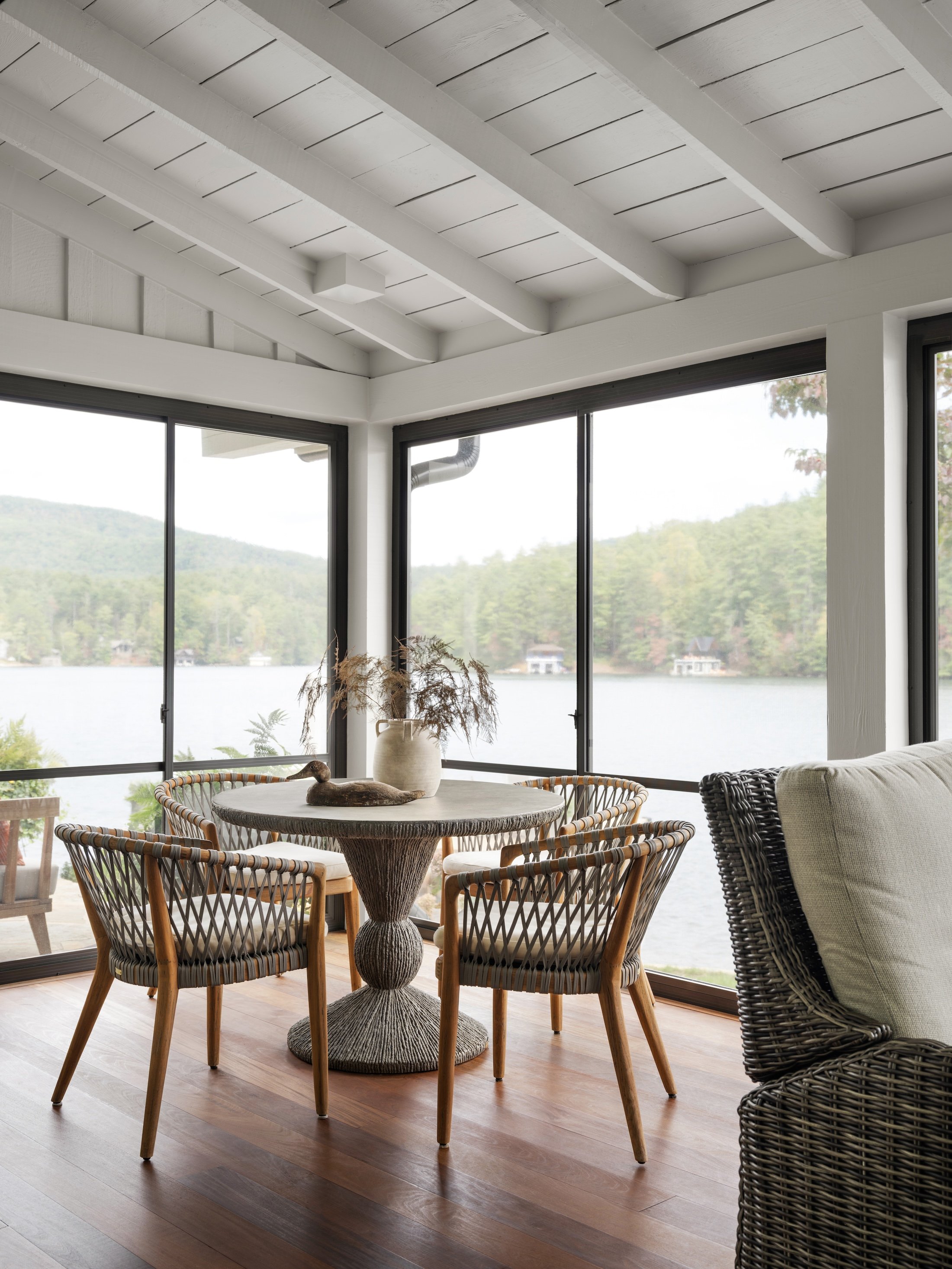 Lakeside dining area with woven chairs, round wood table, white beamed ceiling, and expansive water views.