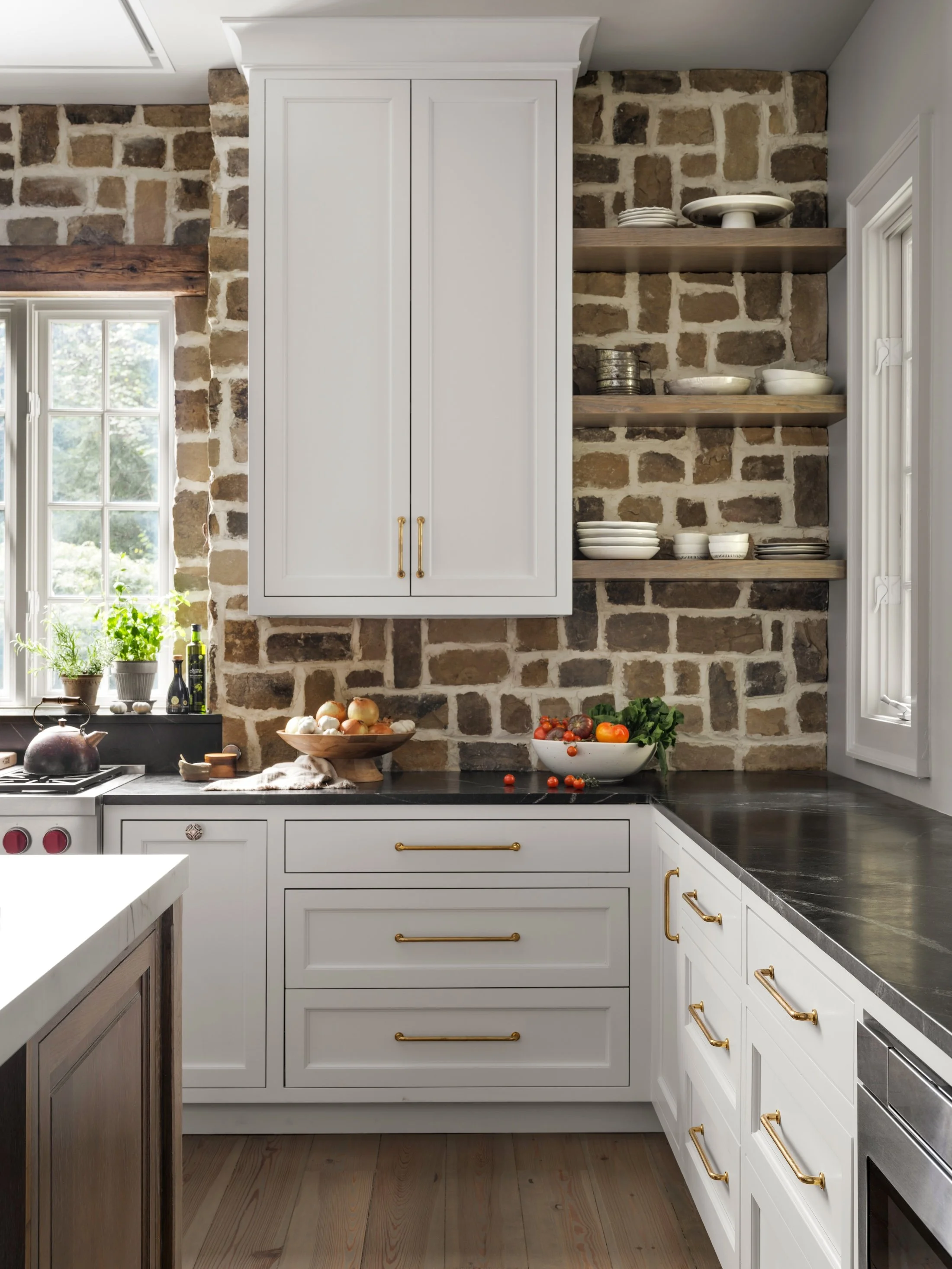 White kitchen with warm wood accents, stone backsplash, black countertops, brass hardware, and layered open shelving.