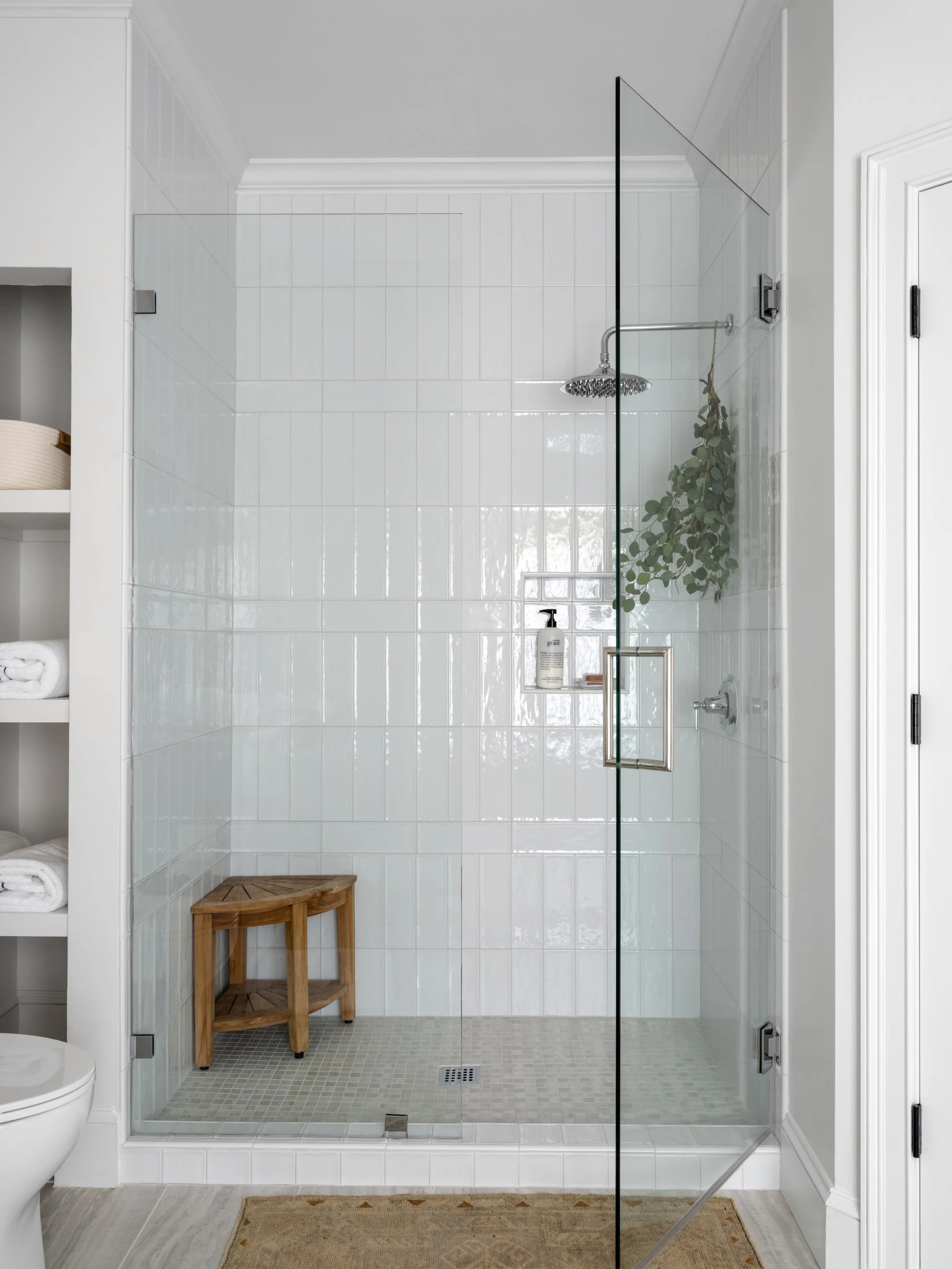 Glass-enclosed shower with glossy white tile, brass fixtures, built-in shelving, and a wooden stool.