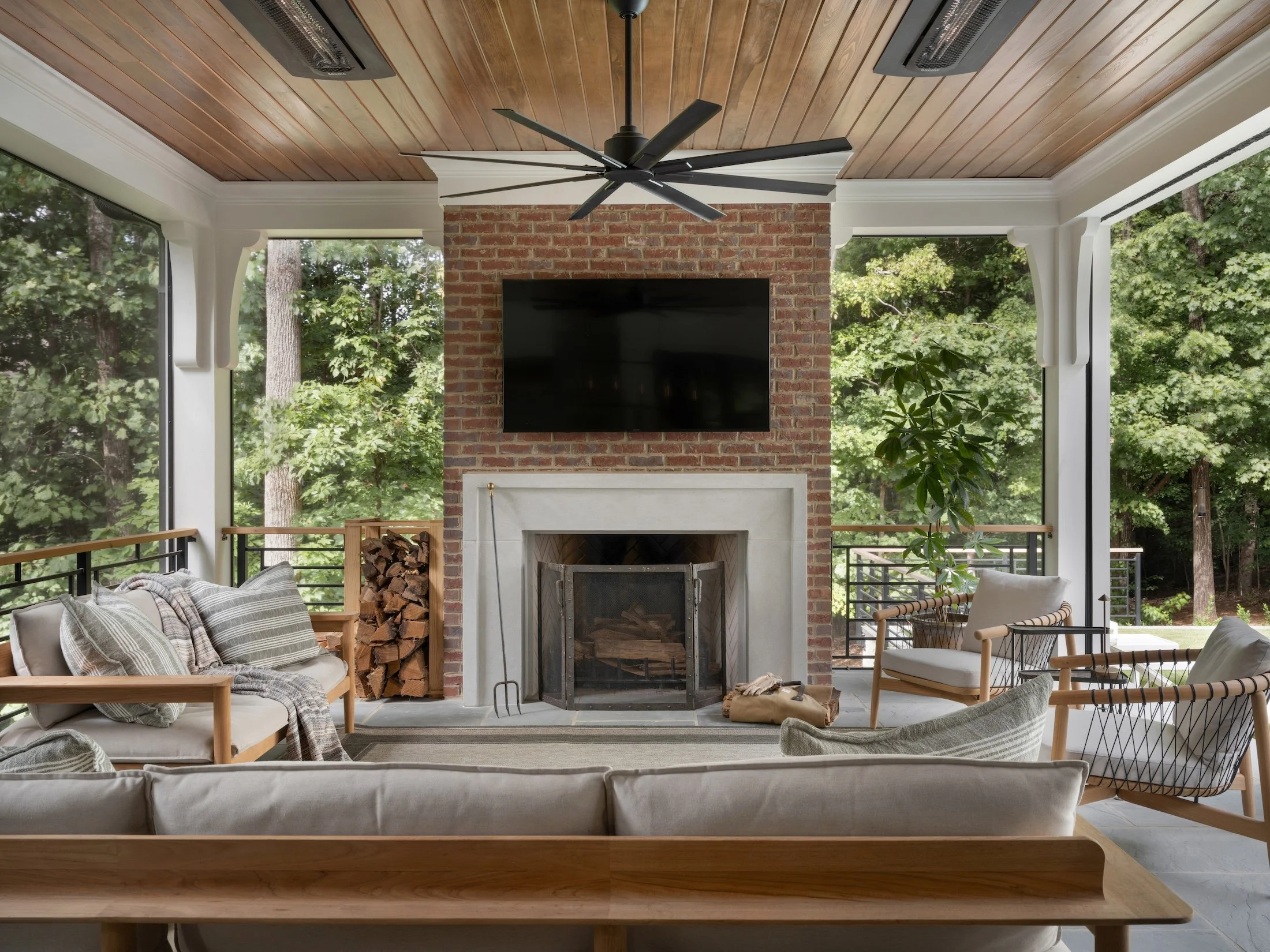Covered outdoor living room with a brick fireplace, wood ceiling, modern seating, and views of the surrounding trees.