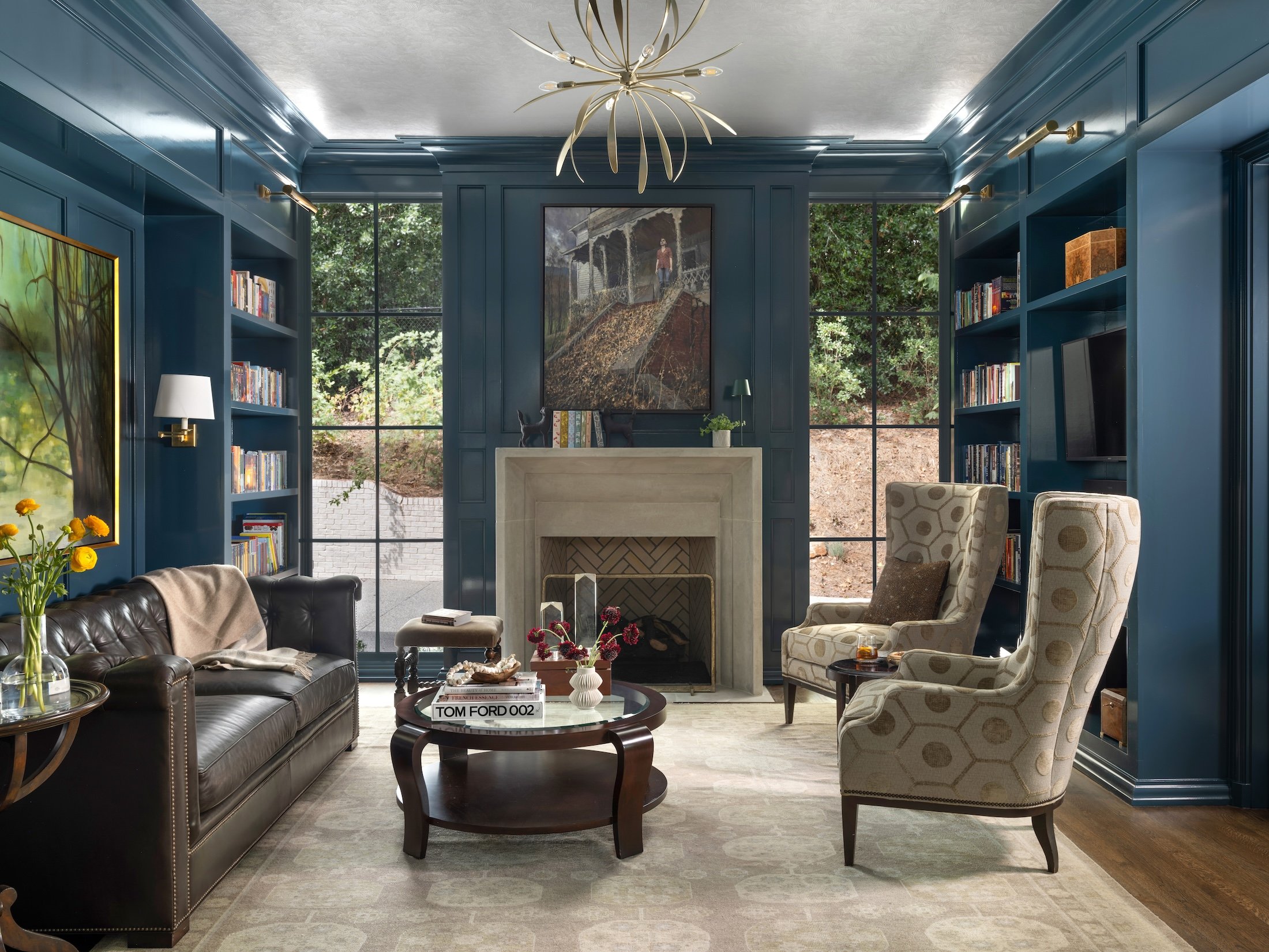 Moody blue library with built-in bookshelves, a stone fireplace, black-framed windows, leather sofa, and patterned wingback chairs.