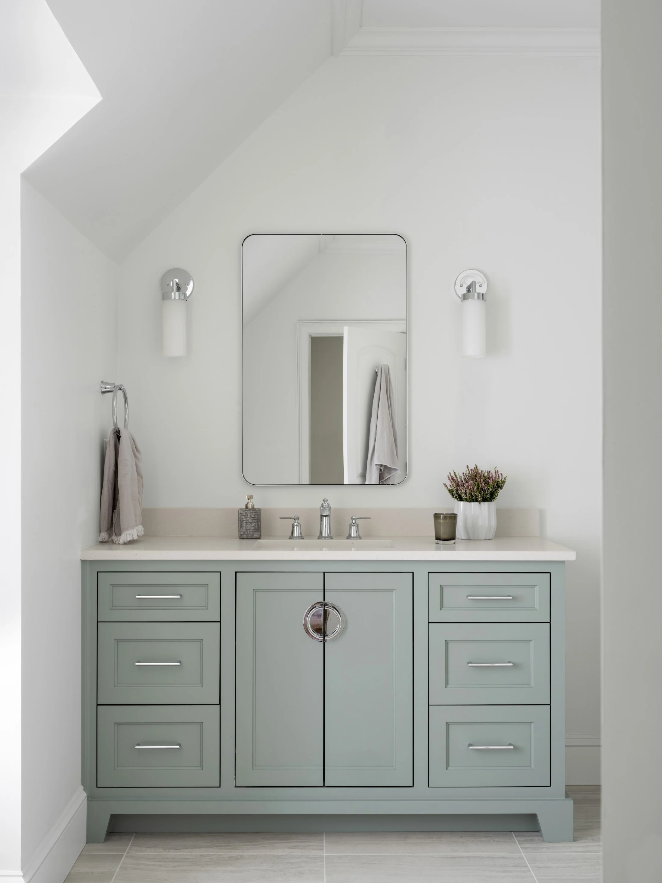 Soft green bathroom vanity with a white countertop, simple mirror, wall sconces, and polished metal fixtures.
