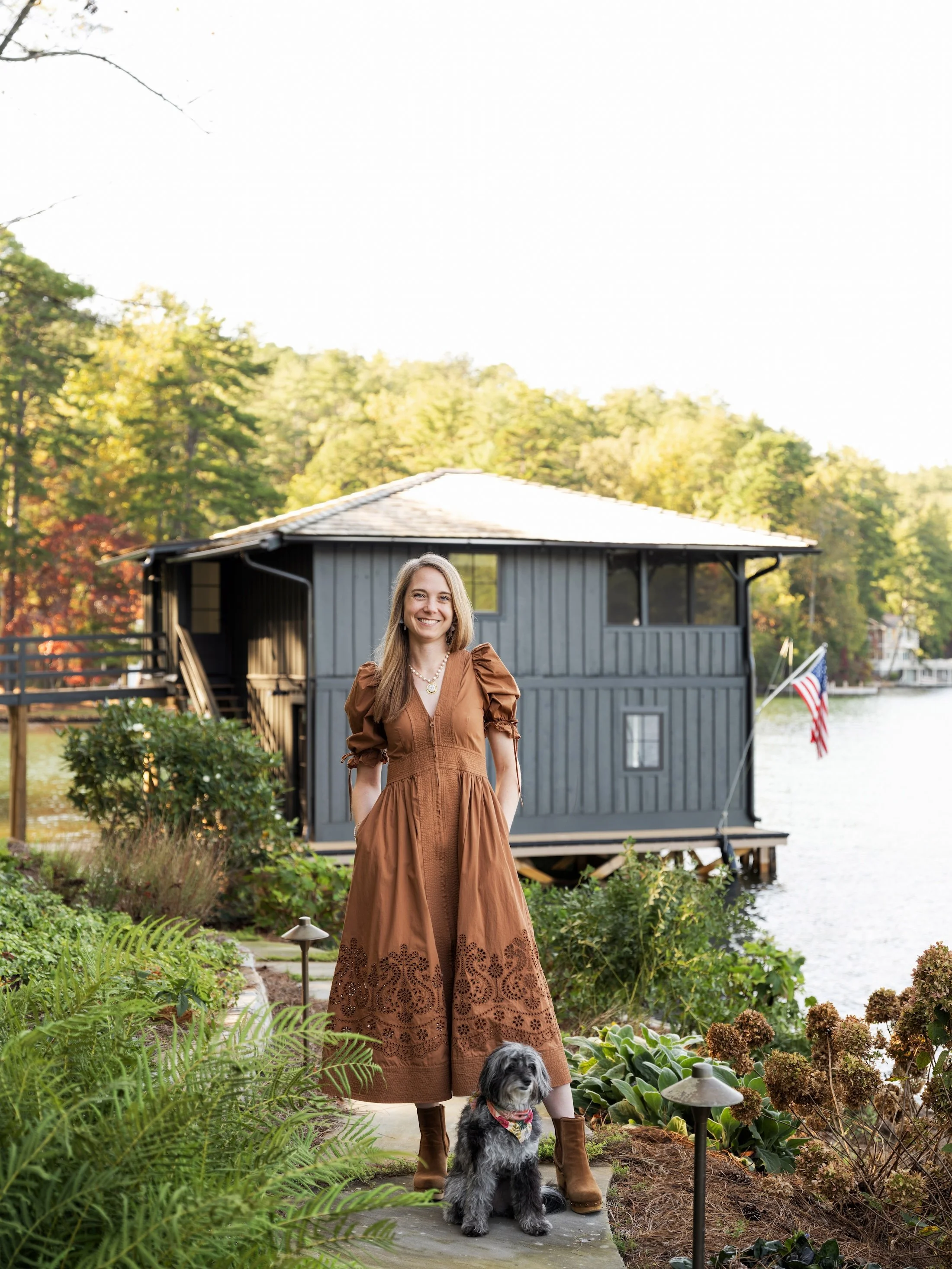 Interior designer Crystal Dalton standing with her dog beside a lakeside boathouse surrounded by lush landscaping.