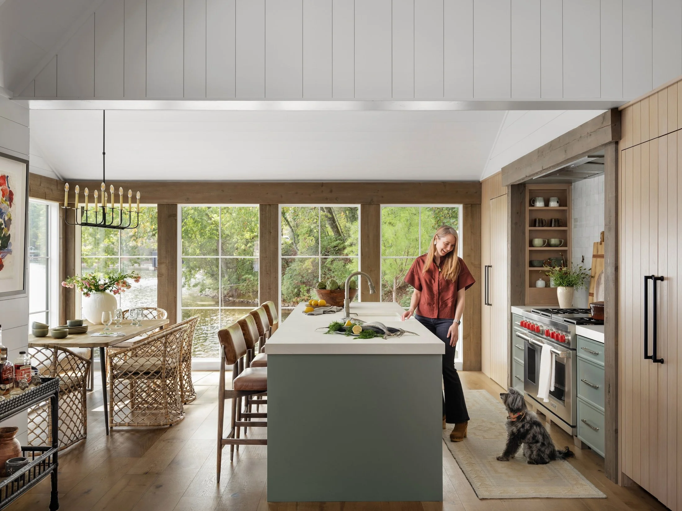 Crystal Dalton standing in a light-filled custom kitchen with her dog beside a sage green island and lakeside views beyond.