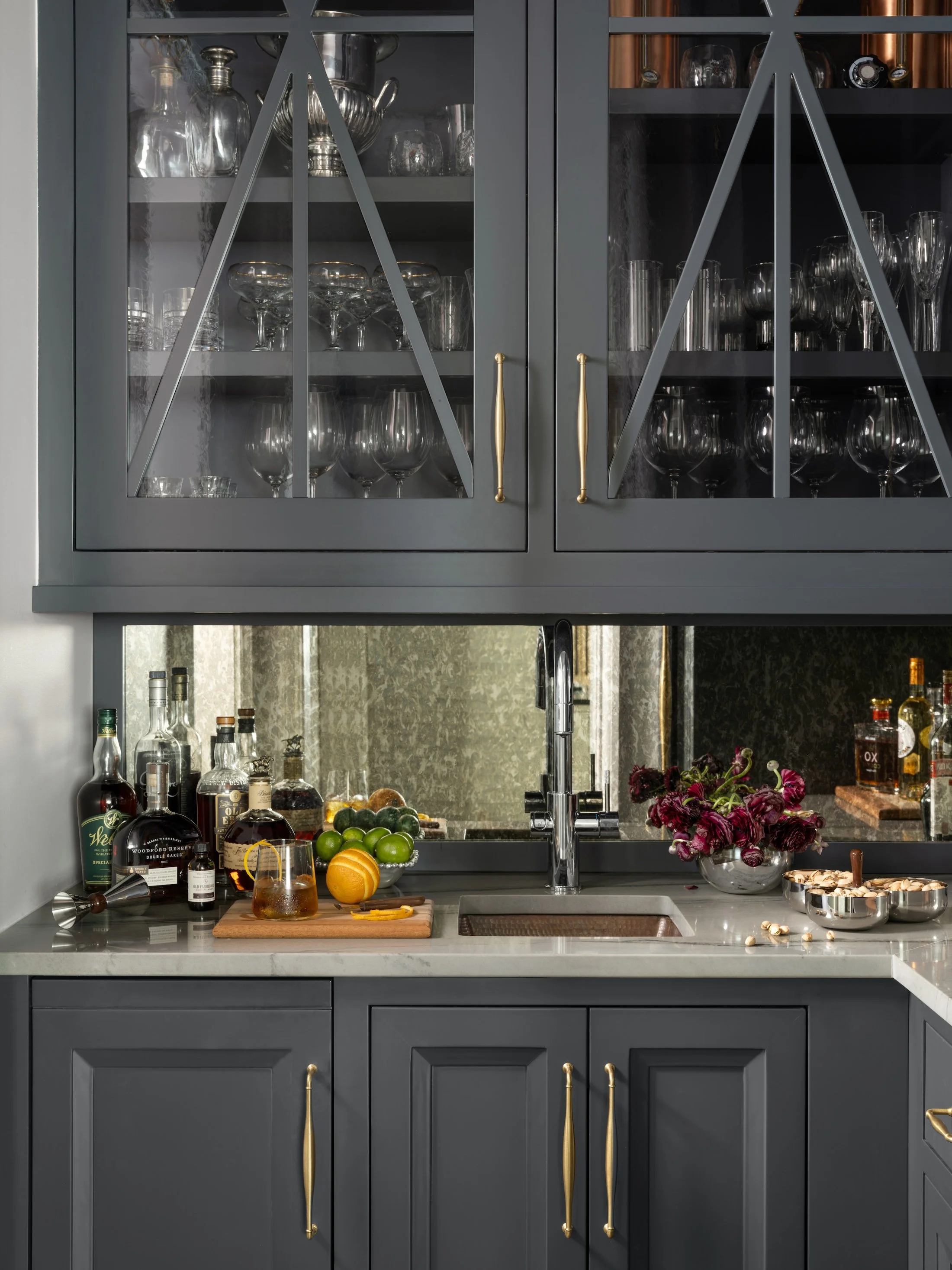 Moody custom home bar with dark cabinetry, glass-front upper cabinets, stone countertop, brass hardware, and mirrored backsplash.