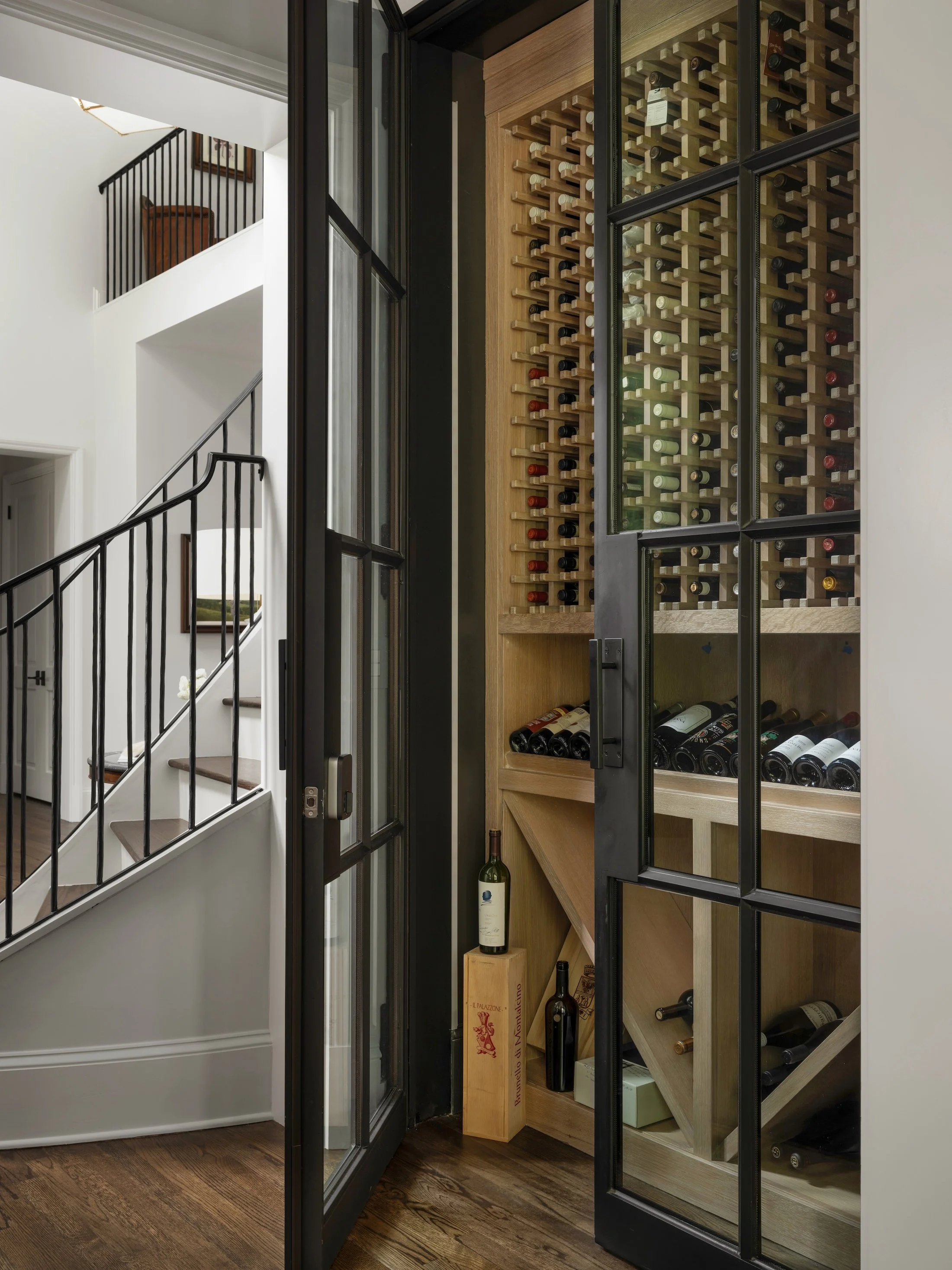 Custom wine cellar with floor-to-ceiling wood bottle storage, black-framed glass door, and adjacent staircase.