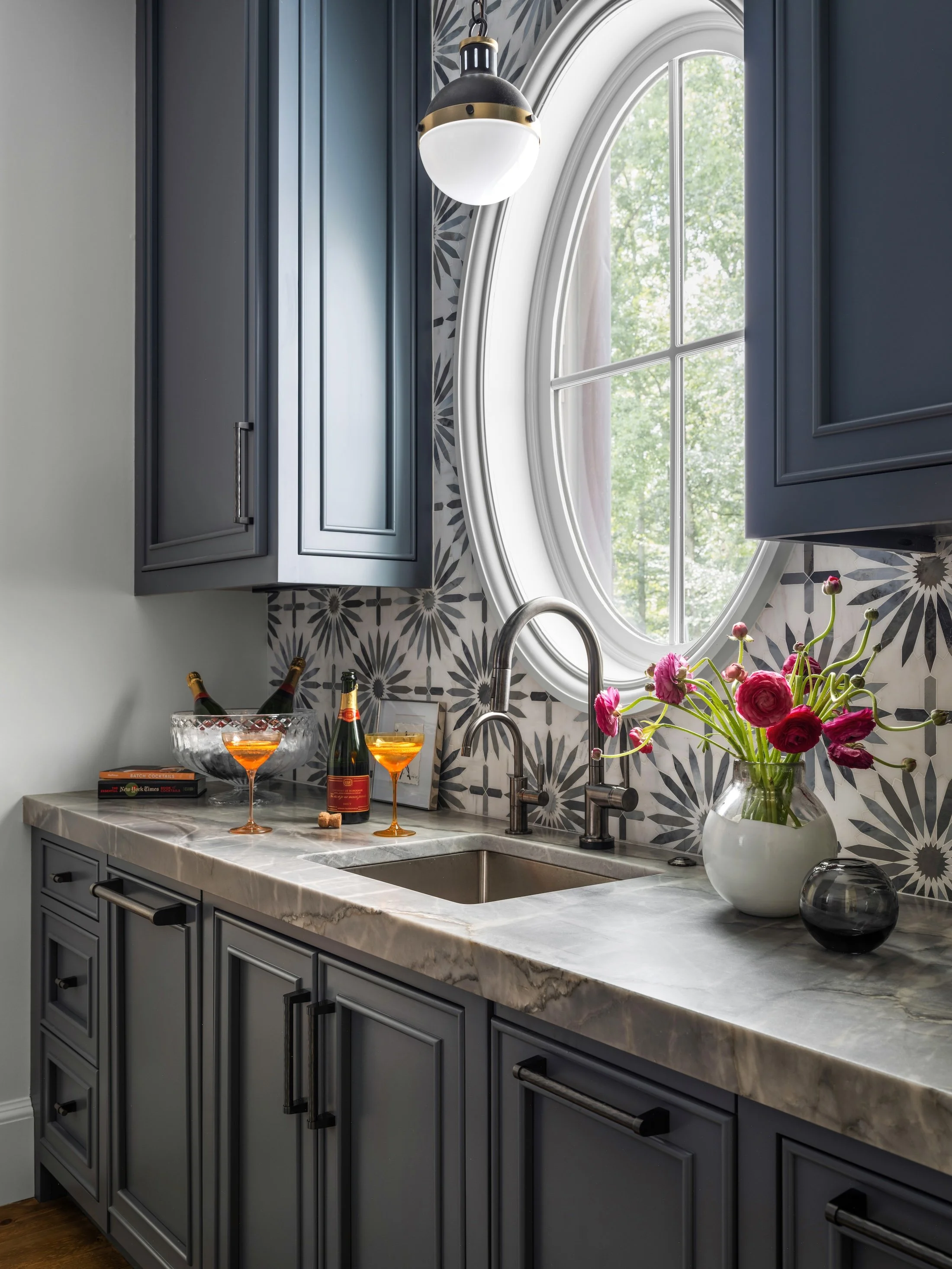 Dark blue wet bar with an arched window, patterned backsplash, stone countertop, brass fixtures, flowers, and cocktail glasses.