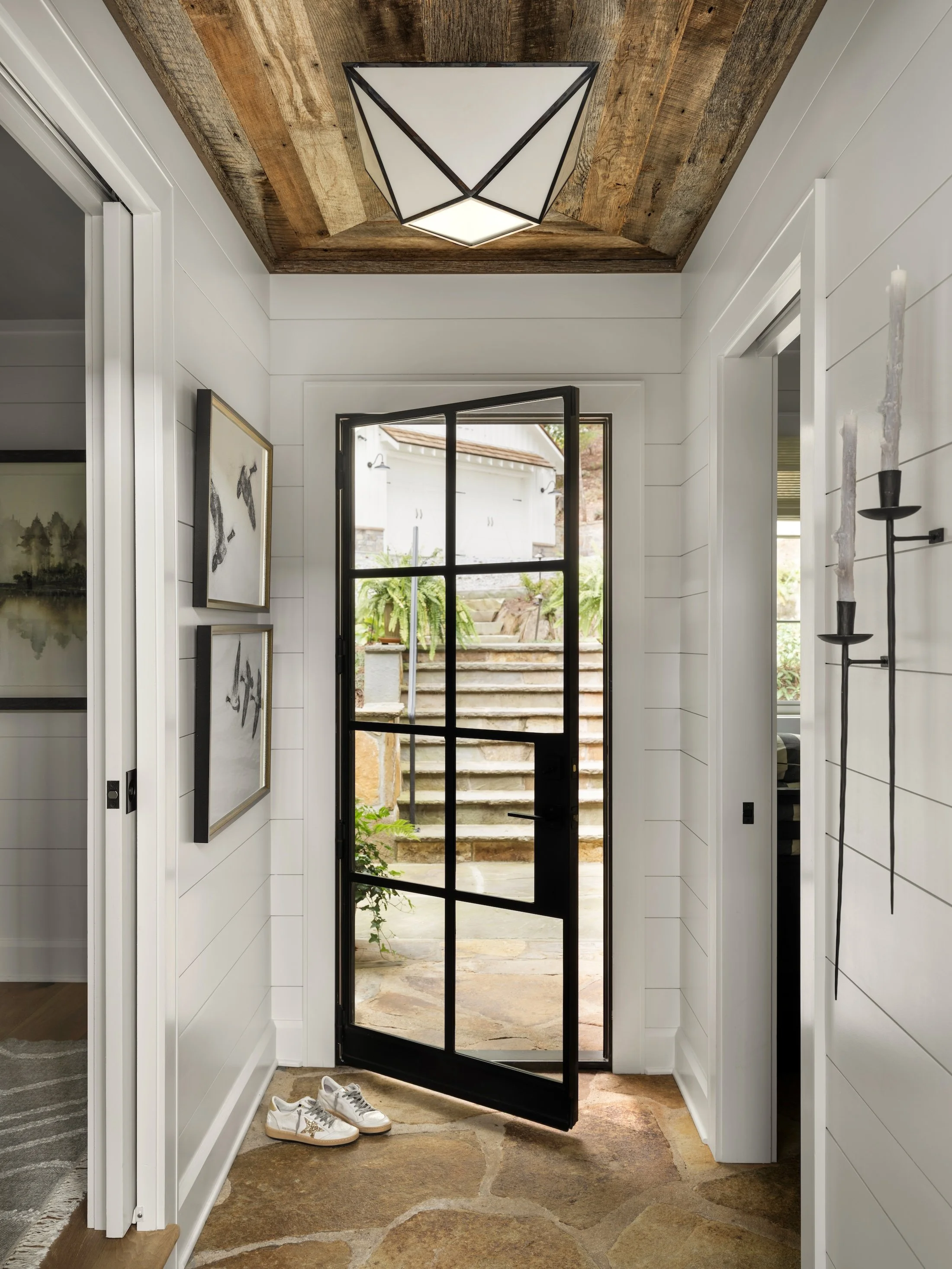 Bright entry hallway with white shiplap walls, black-framed glass door, artwork, and exposed wood ceiling detail.