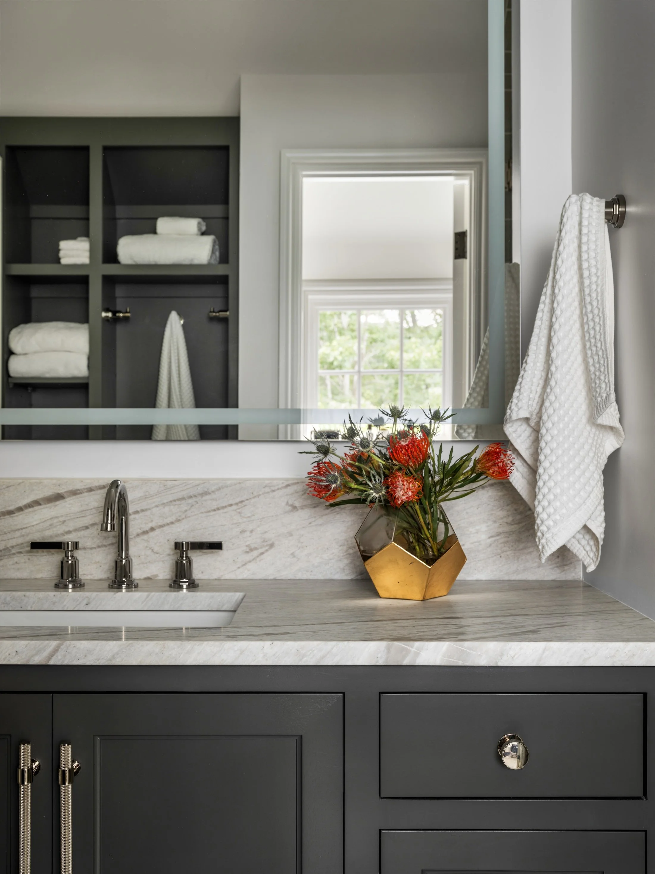 Dark bathroom vanity with marble countertop, built-in linen shelving, large mirror, and colorful floral arrangement.