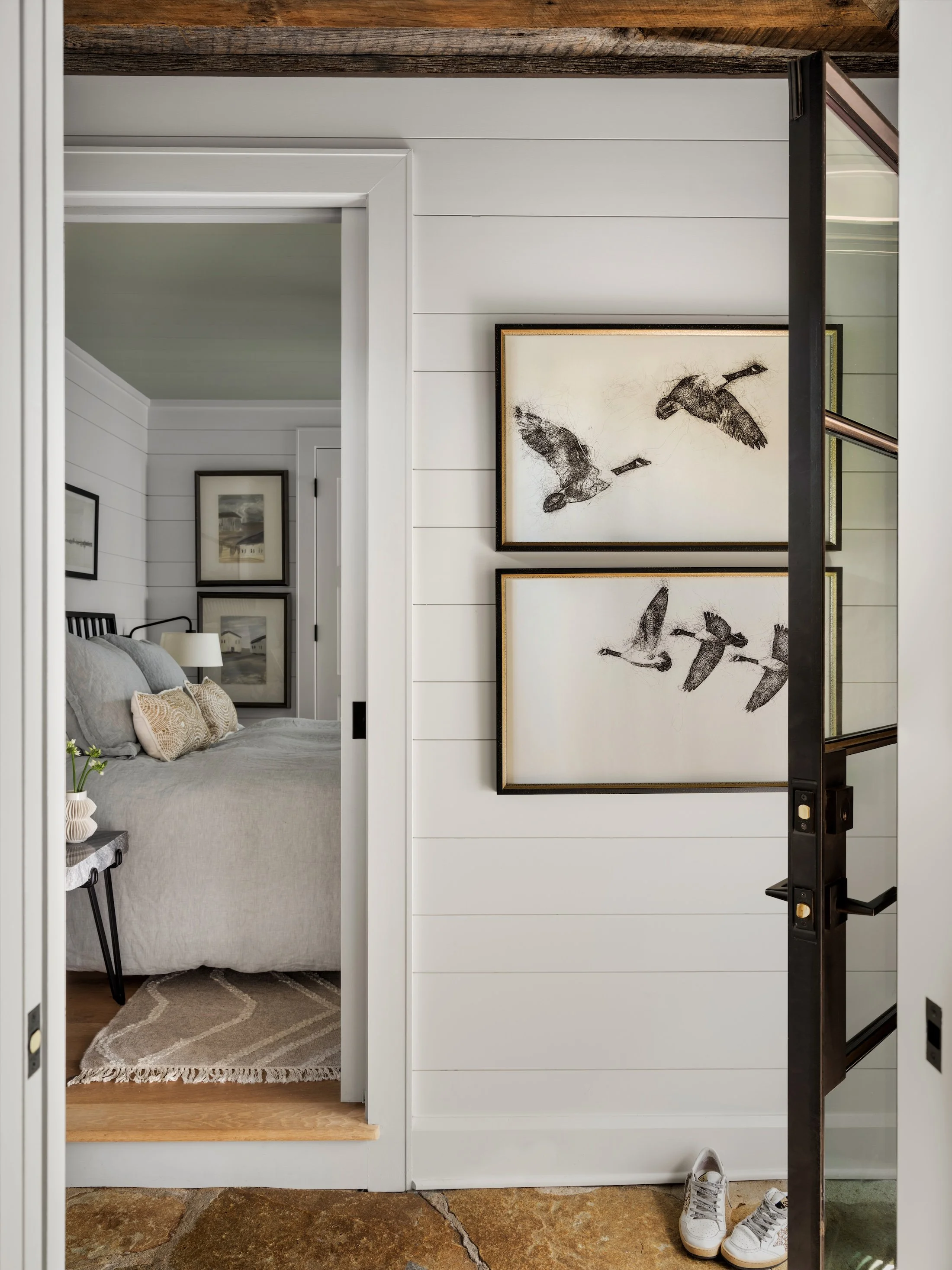 Bedroom entry with white shiplap walls, black-framed glass door, framed artwork, and a glimpse into the adjoining bedroom.