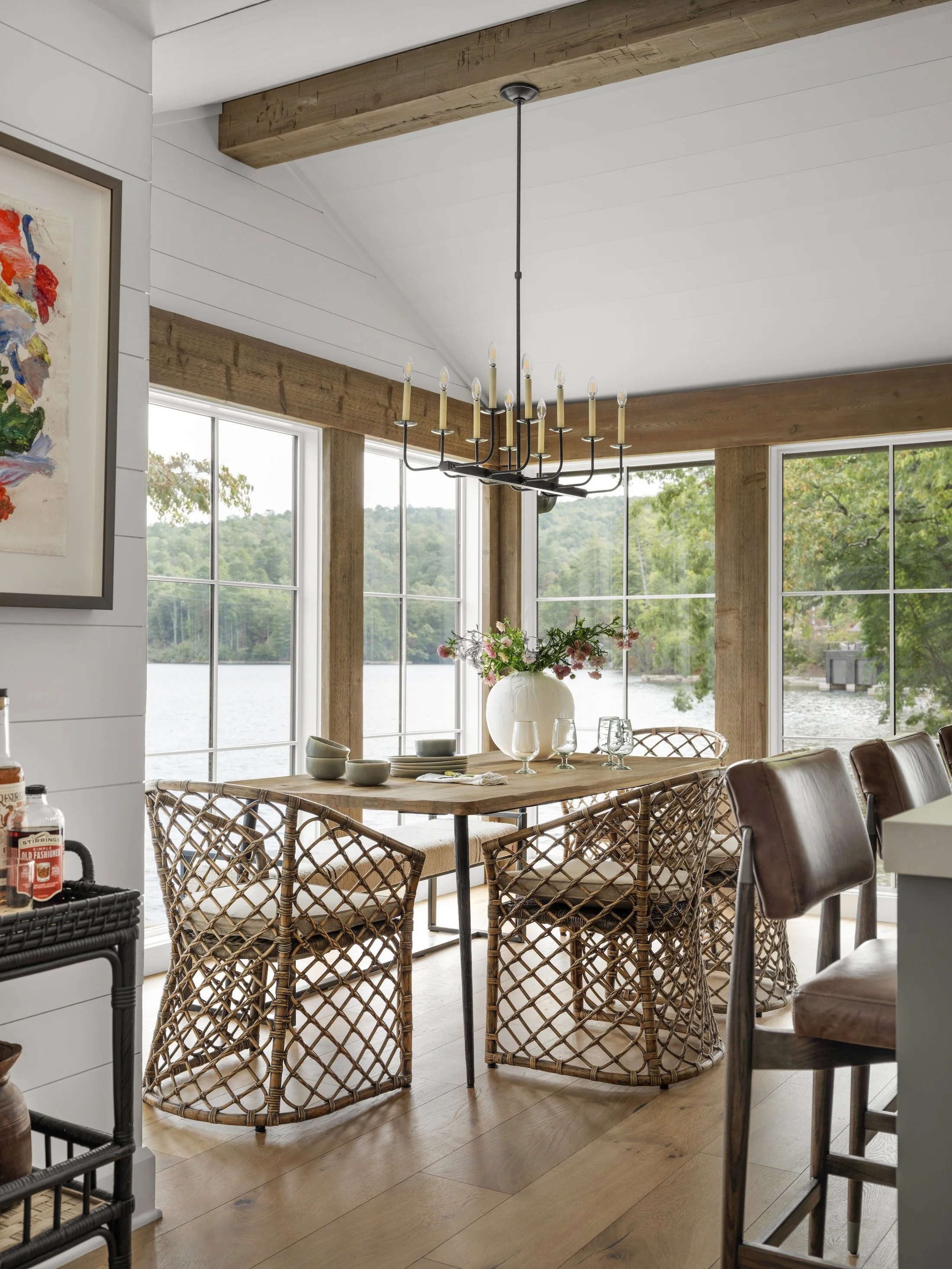 Light-filled dining area with woven chairs, wood table, black chandelier, exposed beams, and large windows overlooking the lake.