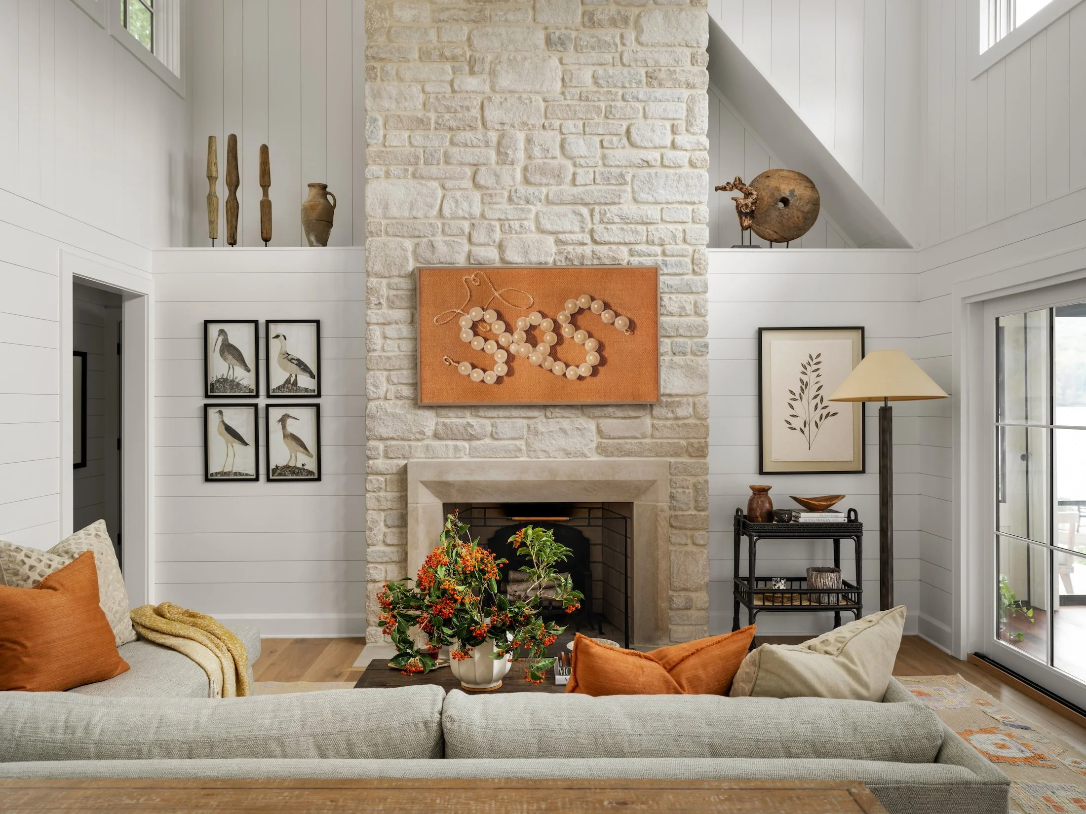 Cozy living room with white shiplap walls, a pale stone fireplace, neutral upholstery, rust-colored pillows, framed artwork, and layered decorative objects.
