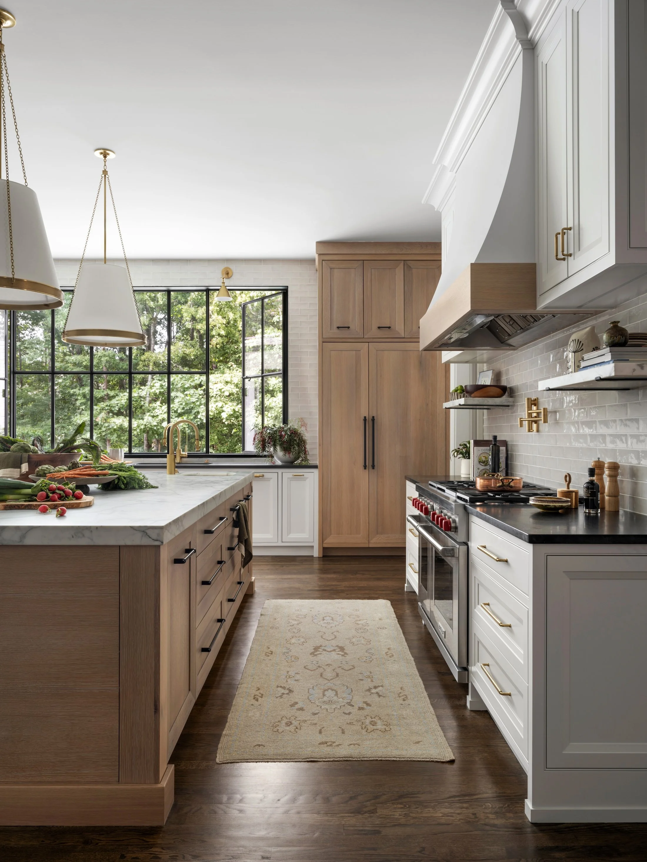 Layered kitchen interior with a natural wood island, white cabinetry, brass fixtures, and large black-framed windows.