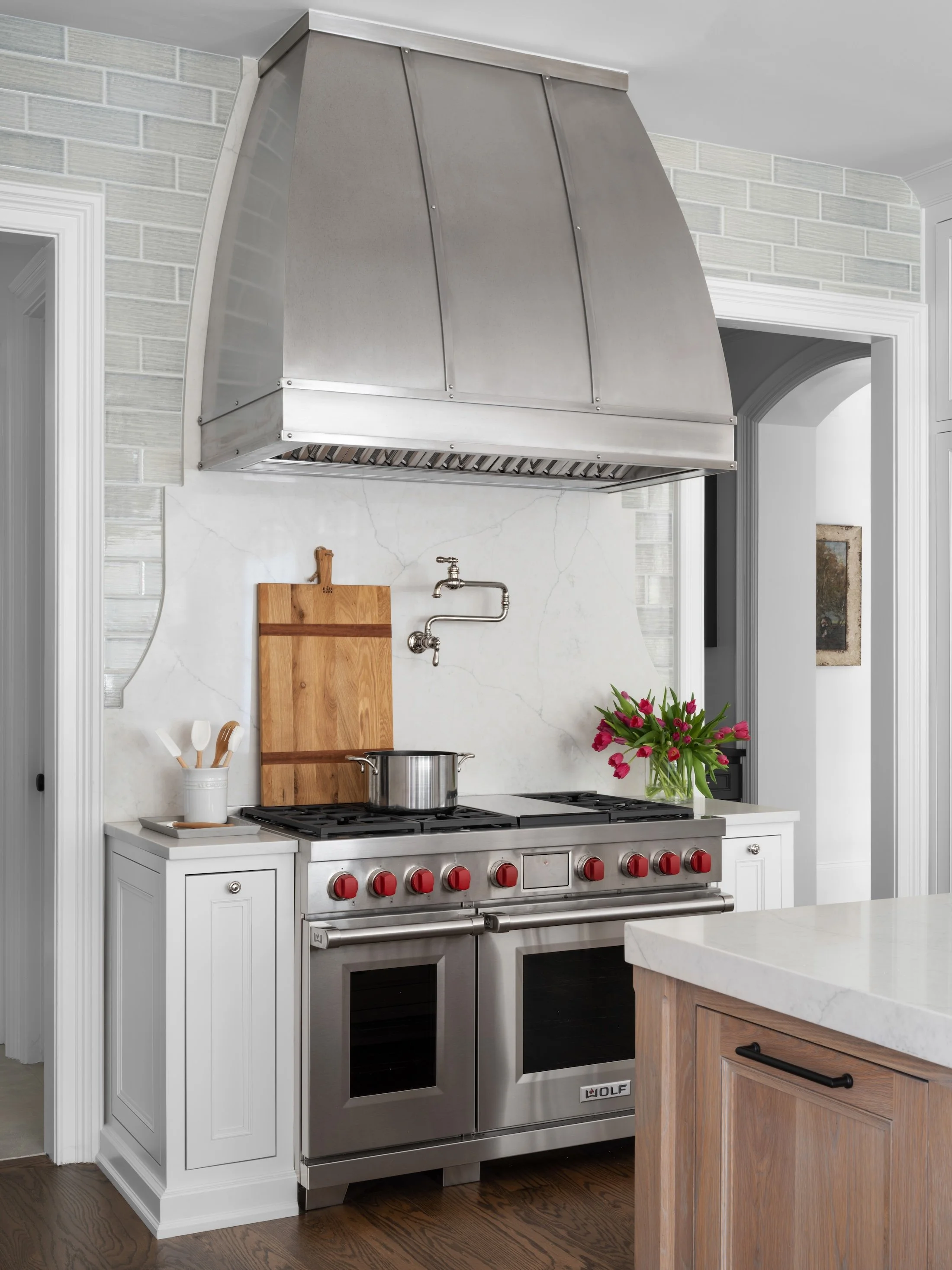White kitchen with a stainless steel range, oversized metal range hood, marble backsplash, and warm wood cabinetry.
