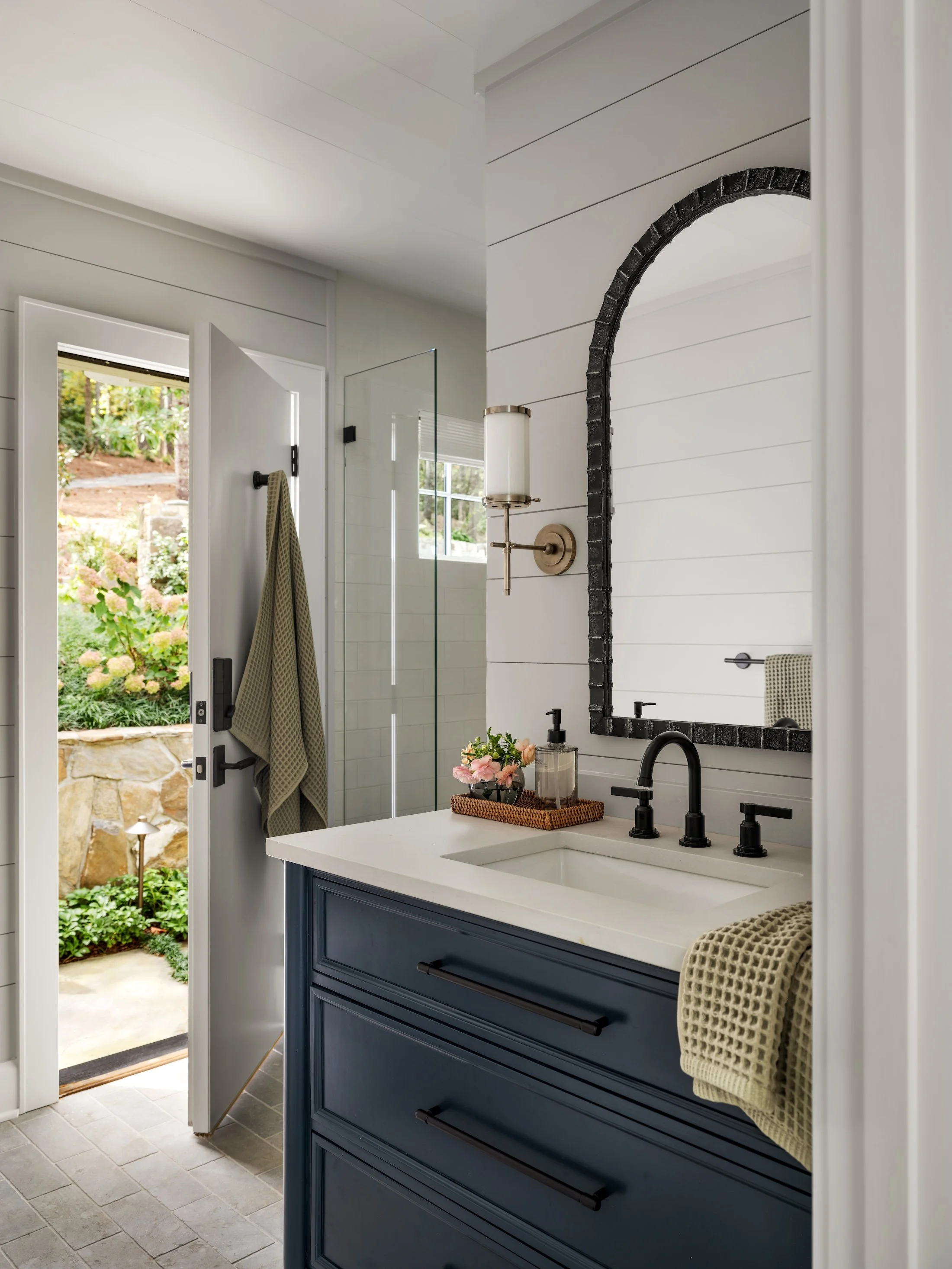 Light-filled bathroom with blue vanity, black fixtures, arched mirror, and an open door leading to the garden.
