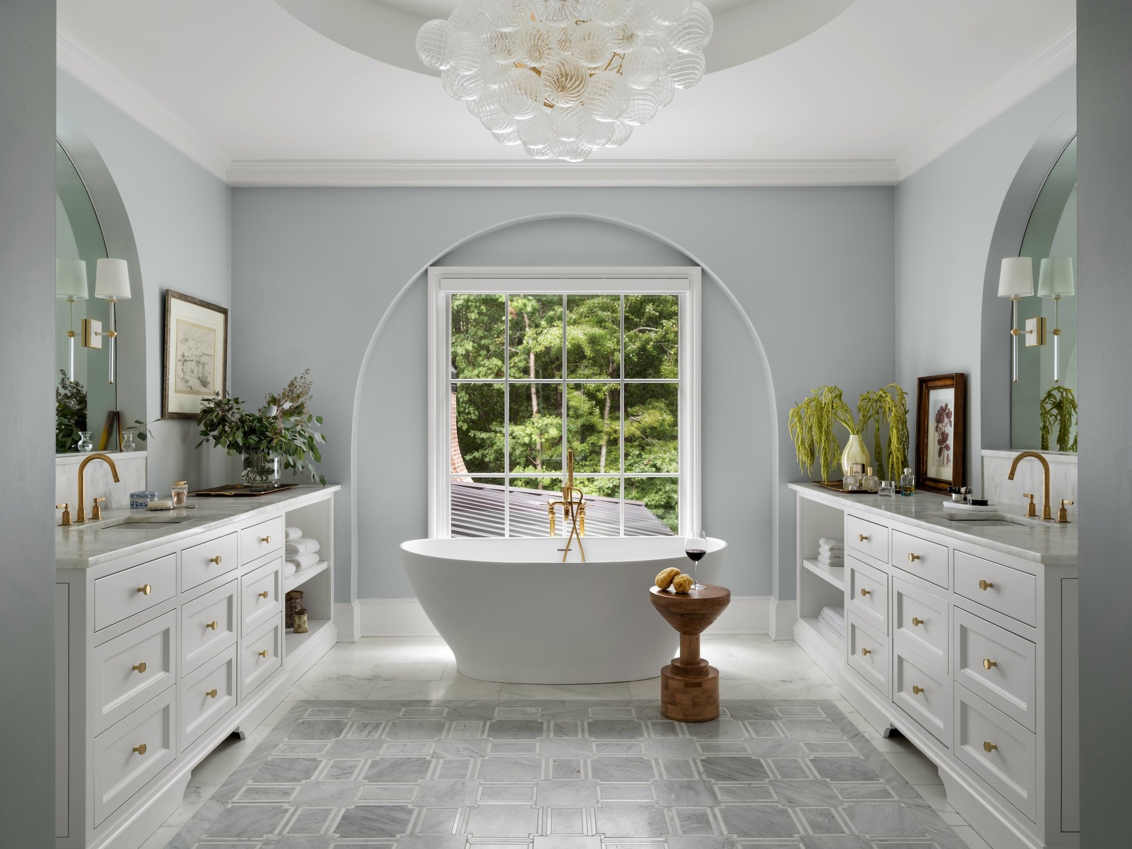 Light blue primary bathroom with a freestanding tub, symmetrical white vanities, brass fixtures, arched architectural detail, and marble flooring.