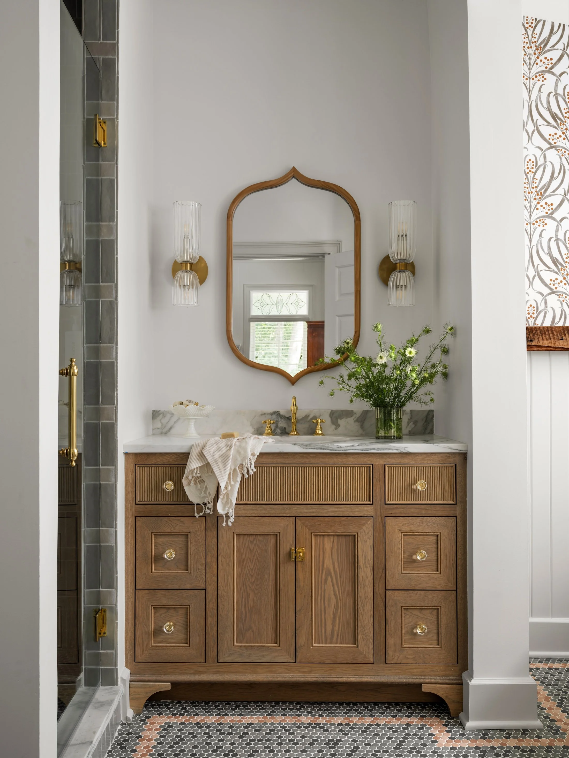 Powder room with an antique wood vanity, decorative mirror, brass fixtures, and patterned wallpaper.