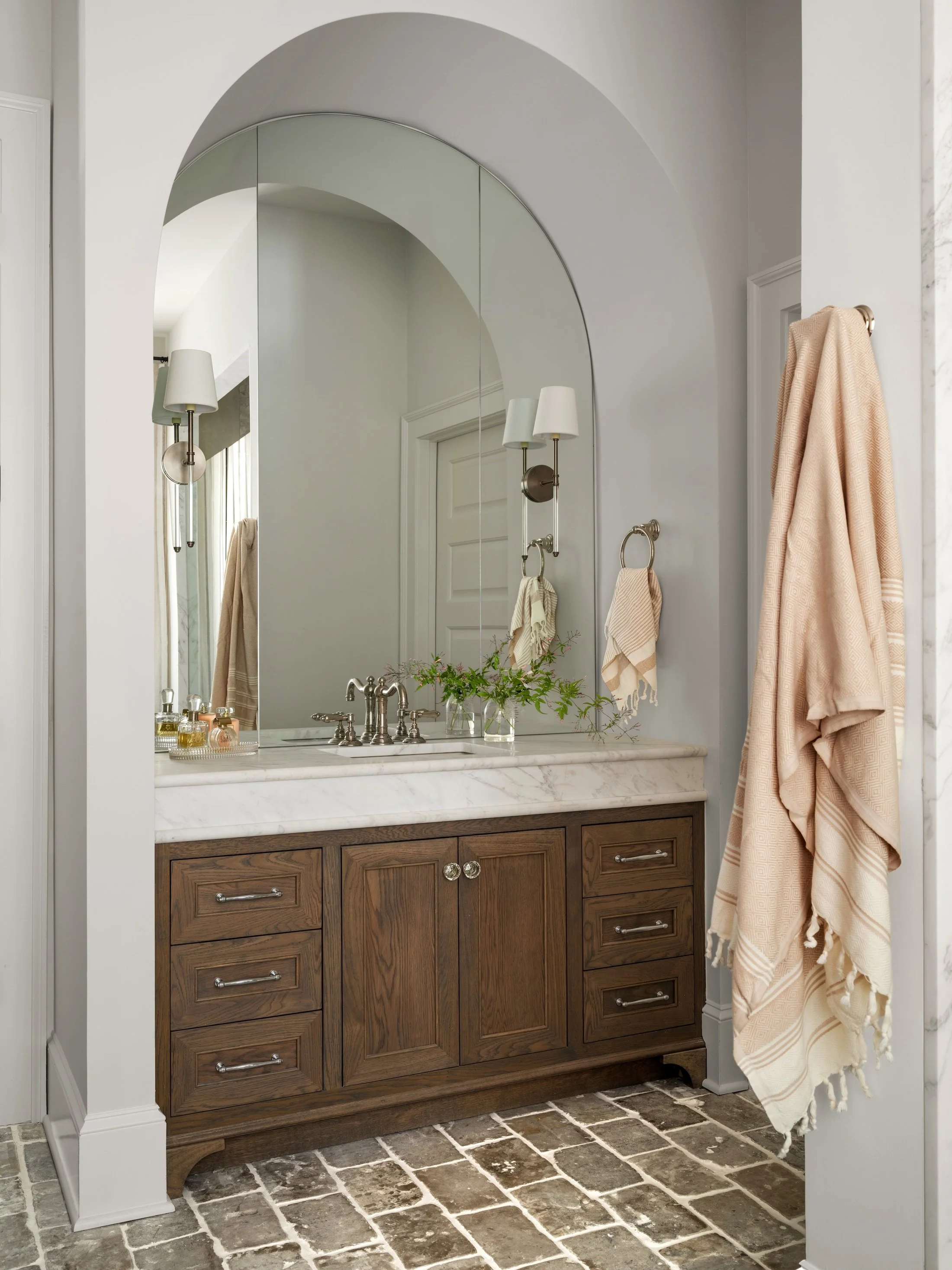 Bathroom with a dark wood vanity, marble countertop, oversized arched mirror, and soft neutral finishes.