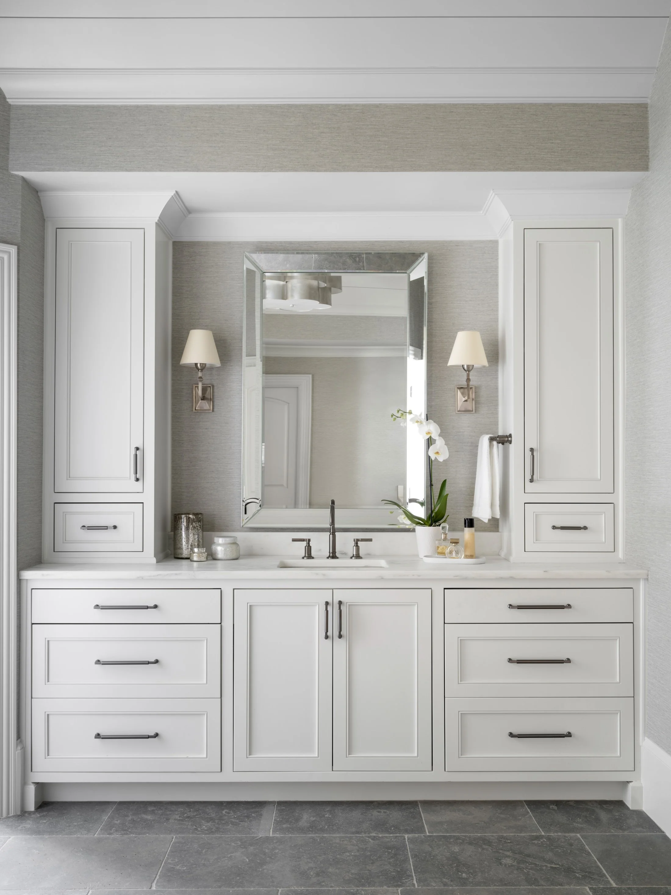 Symmetrical white bathroom vanity with a large framed mirror, stone countertop, wall sconces, and built-in cabinetry.