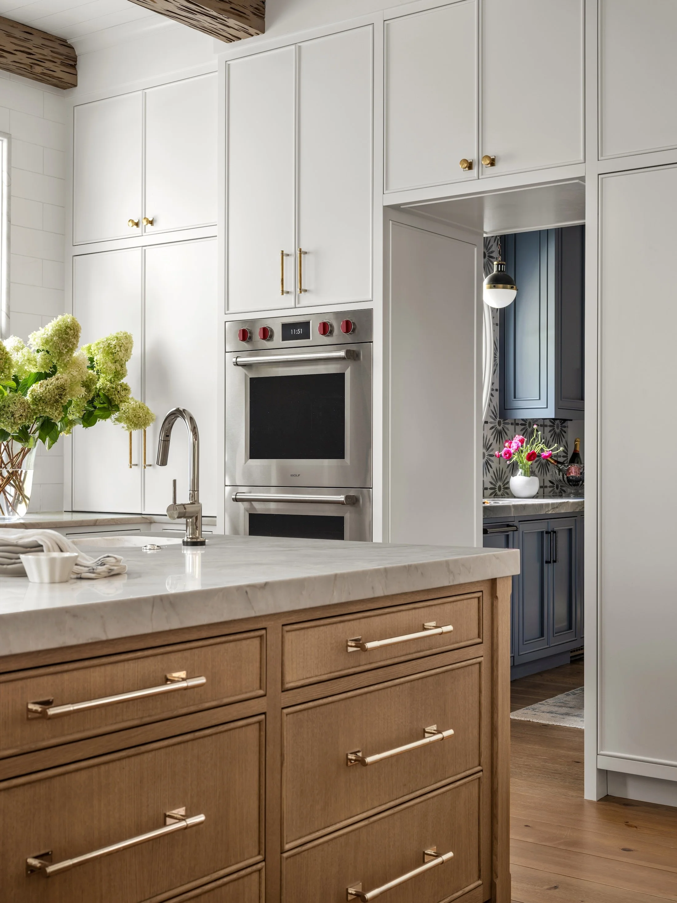 Custom kitchen with white cabinetry, natural wood drawers, marble countertops, brass hardware, and built-in appliances.
