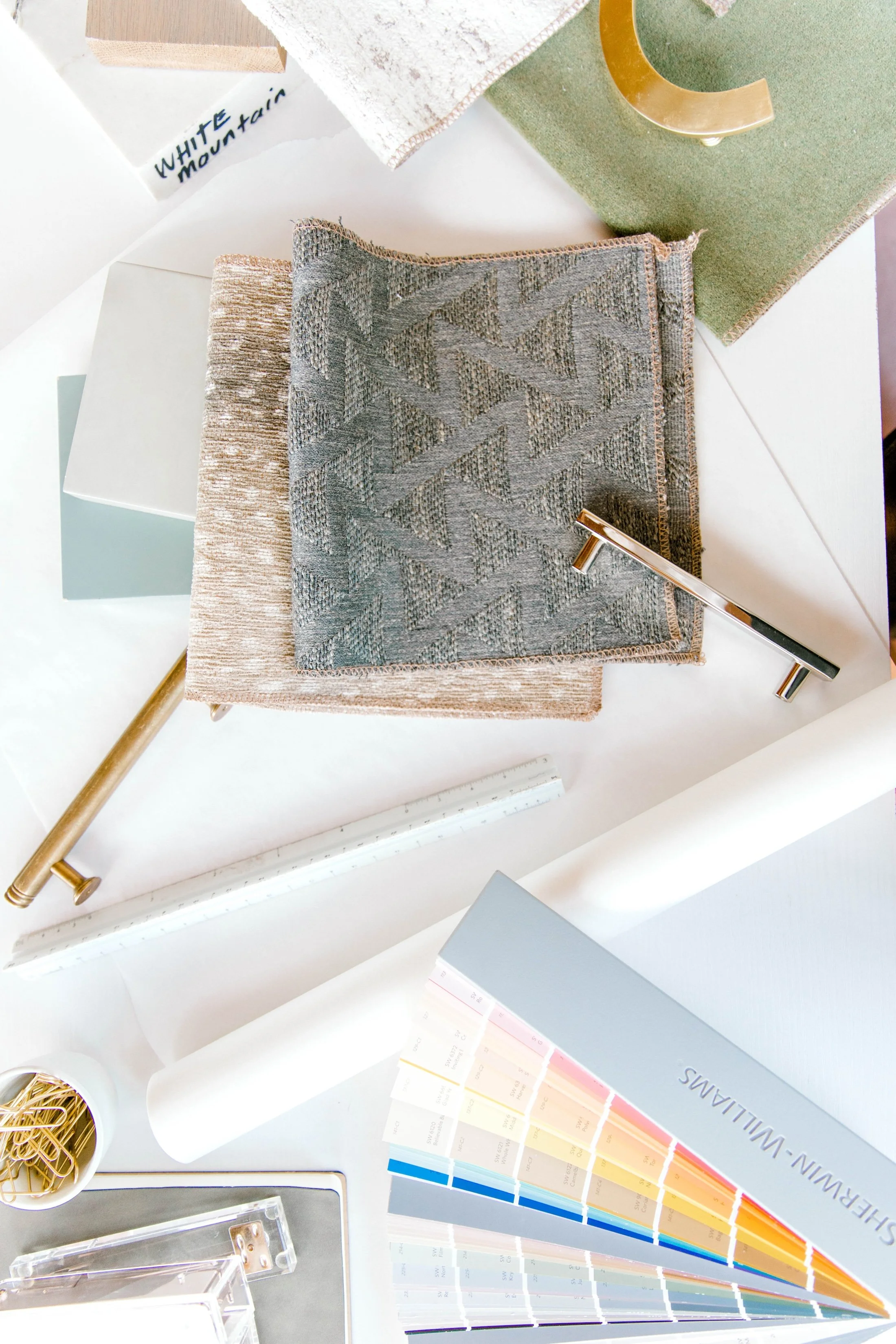 Interior design material flat lay with layered fabric swatches, paint samples, brass hardware, ruler, and color deck.