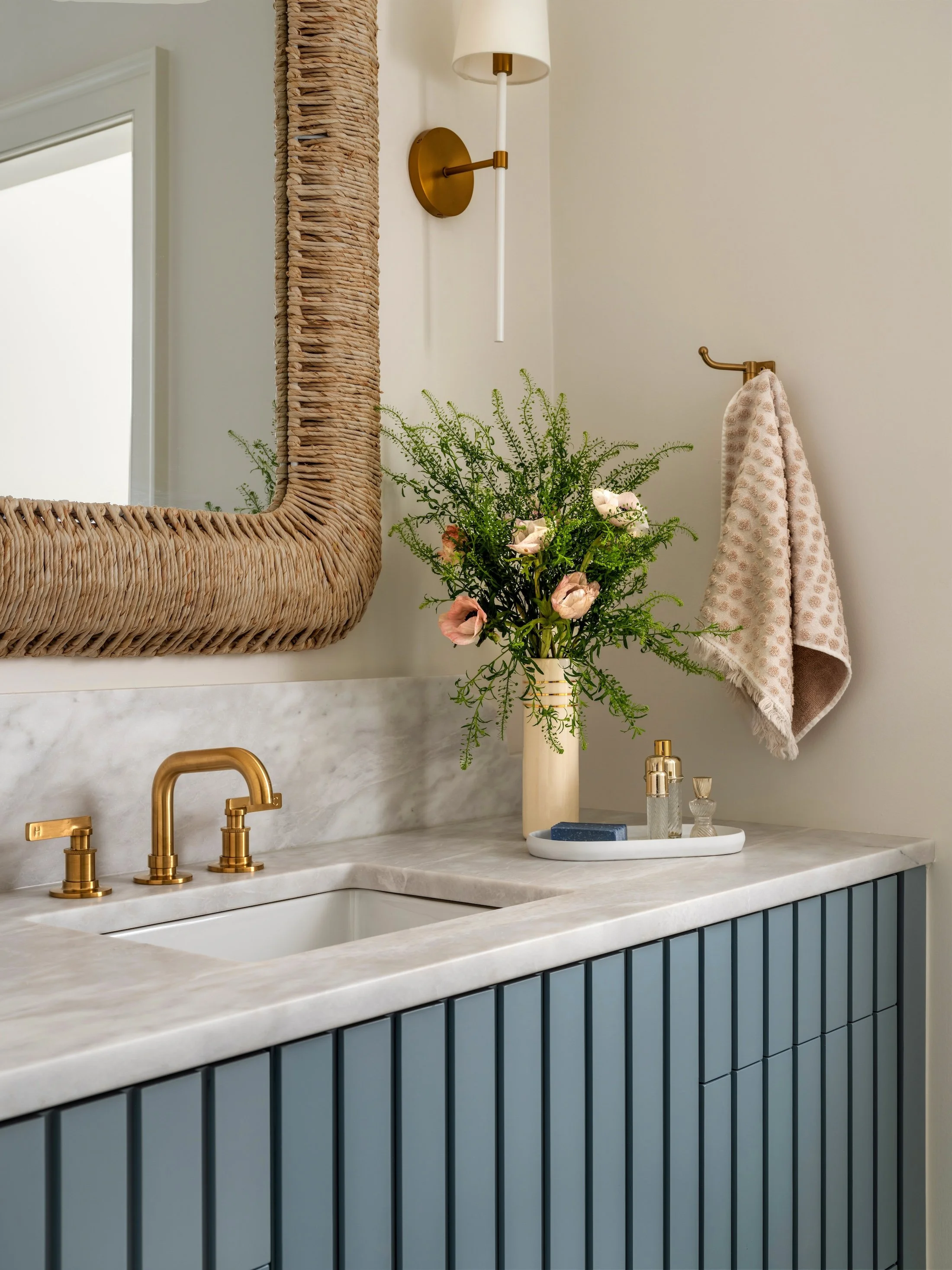 Blue fluted bathroom vanity with marble countertop, brass faucet, woven mirror, fresh flowers, and softly patterned textiles.
