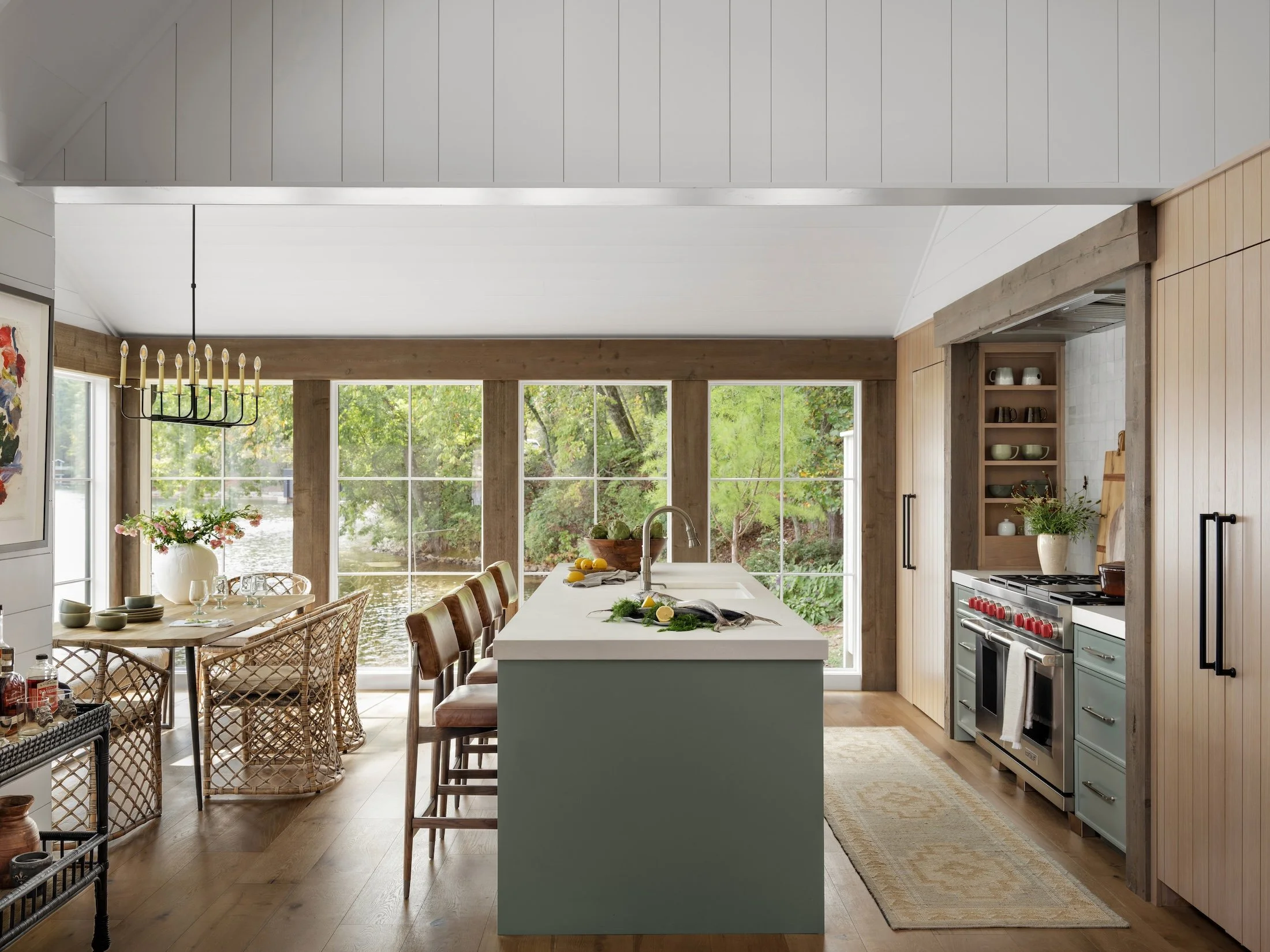 Light-filled kitchen and dining area with a sage green island, woven dining chairs, wood cabinetry, stainless steel range, and expansive windows overlooking the water.