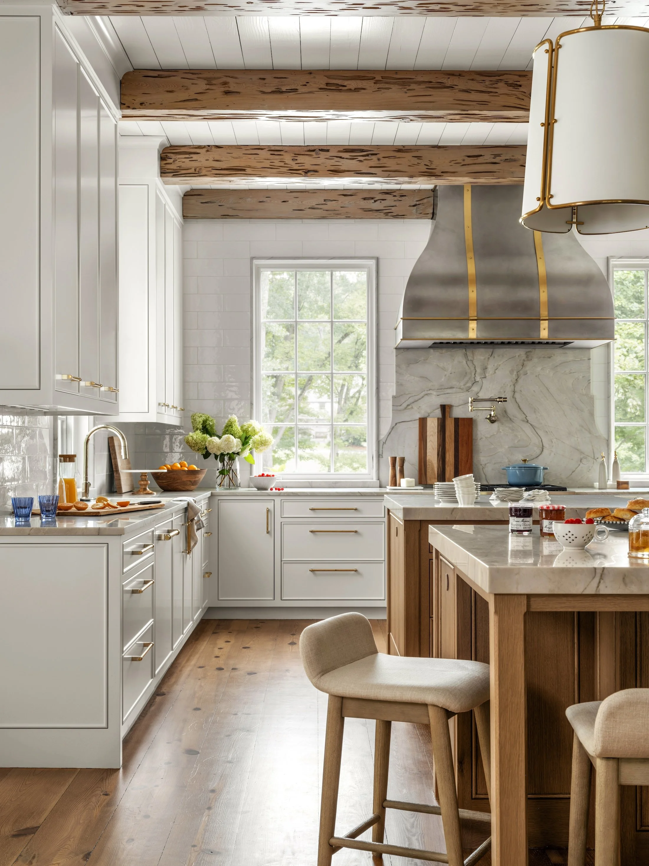Bright white kitchen with exposed wood ceiling beams, custom cabinetry, brass range hood, and a natural wood island.