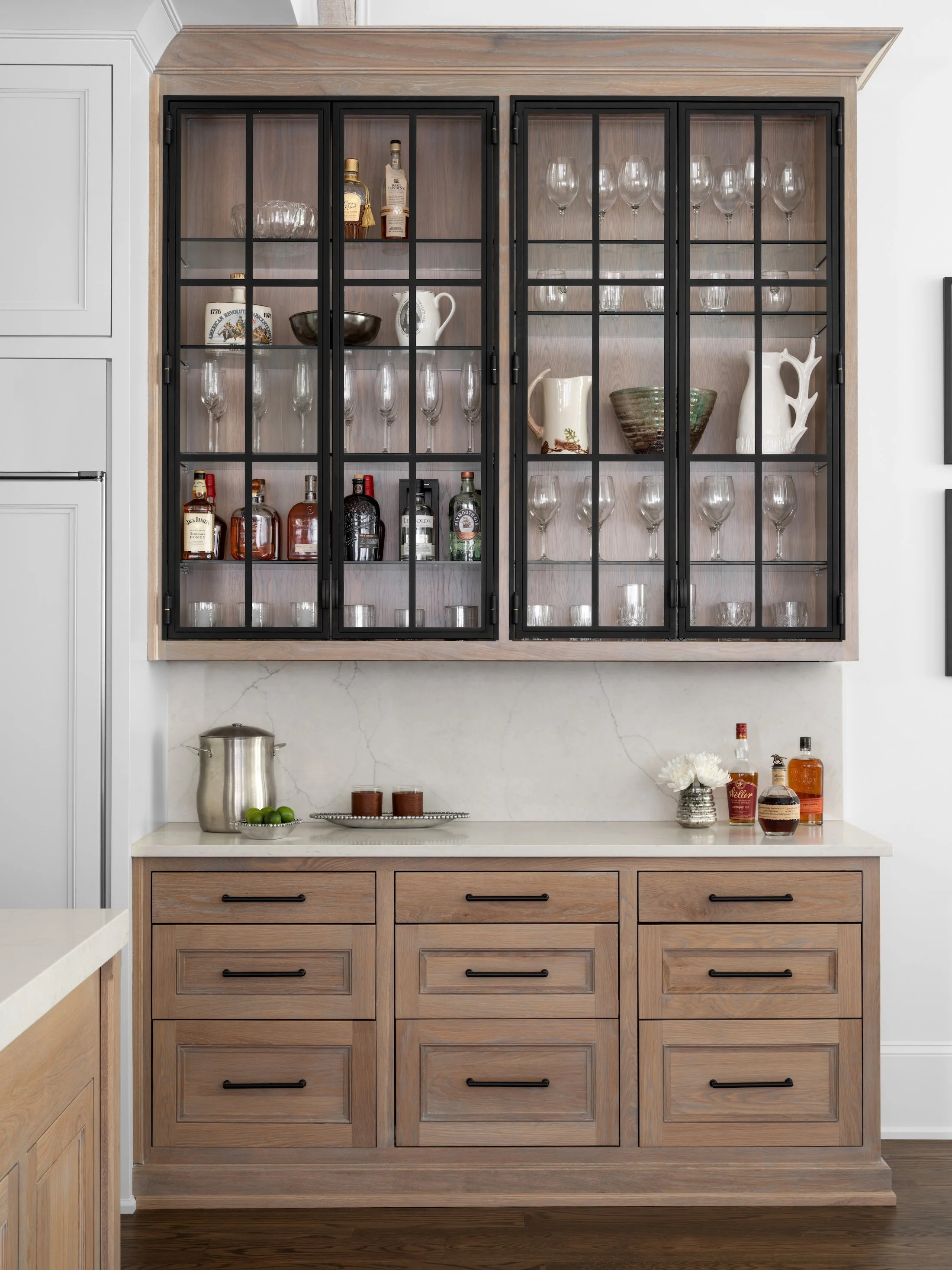 Custom wet bar with glass-front upper cabinets, natural wood drawers, stone countertop, and styled glassware.