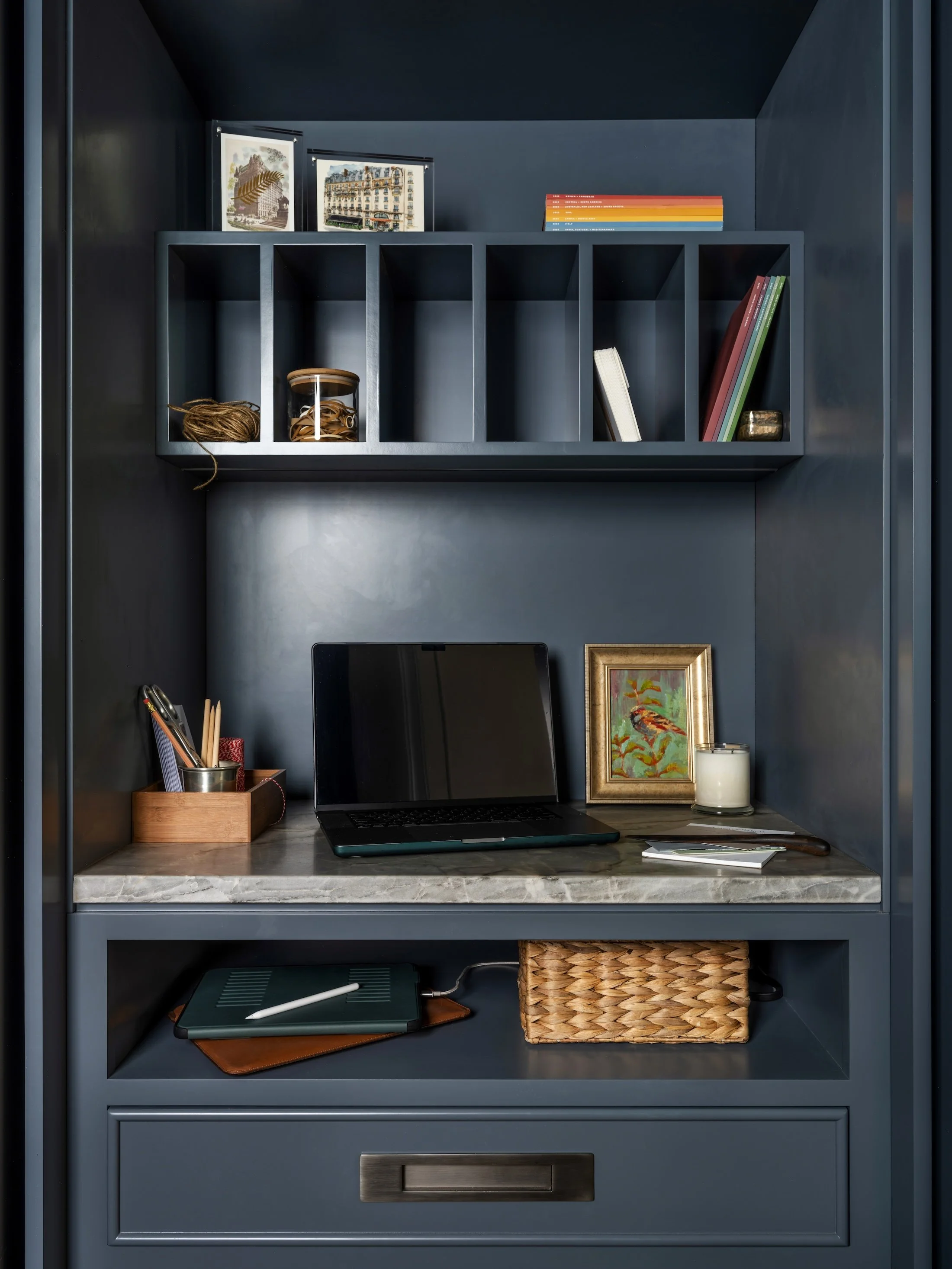 Dark blue built-in desk nook with open shelving, stone desktop, laptop, books, and woven storage baskets.
