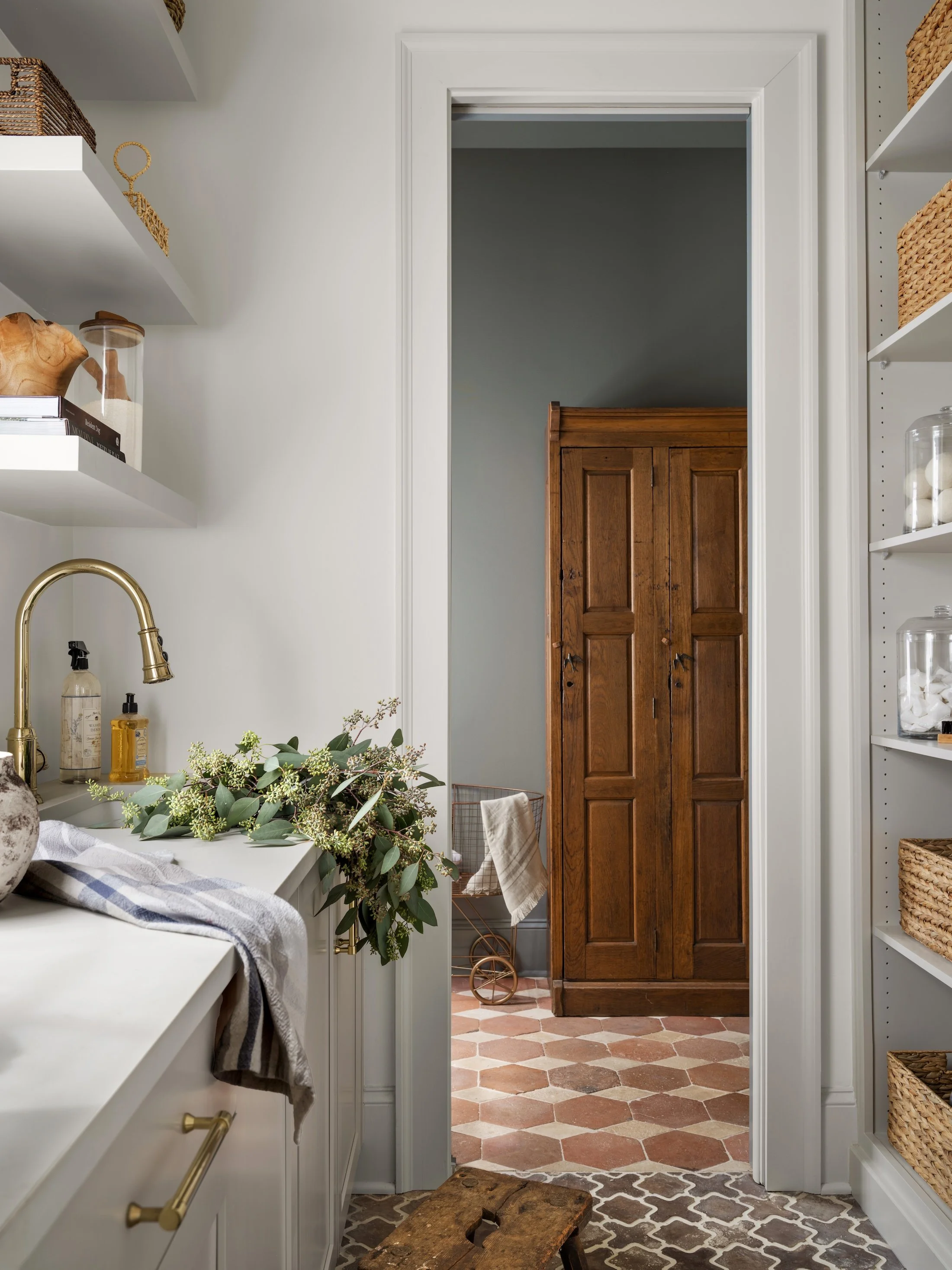White utility sink with brass faucet, open shelving, fresh greenery, and a view into a hallway with patterned tile and an antique wood cabinet.
