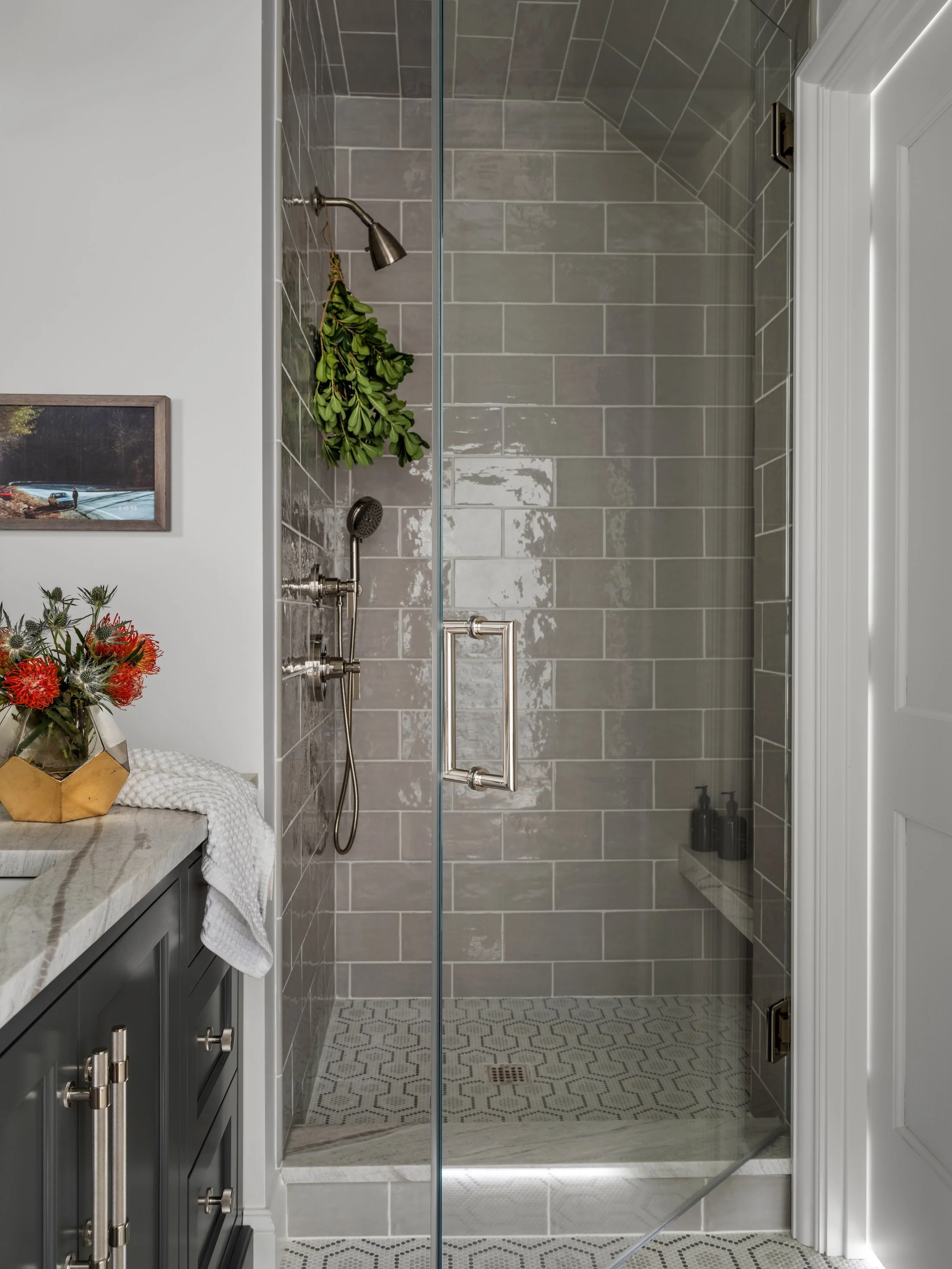 Glass-enclosed shower with glossy gray tile, brass fixtures, and a pebble tile floor.