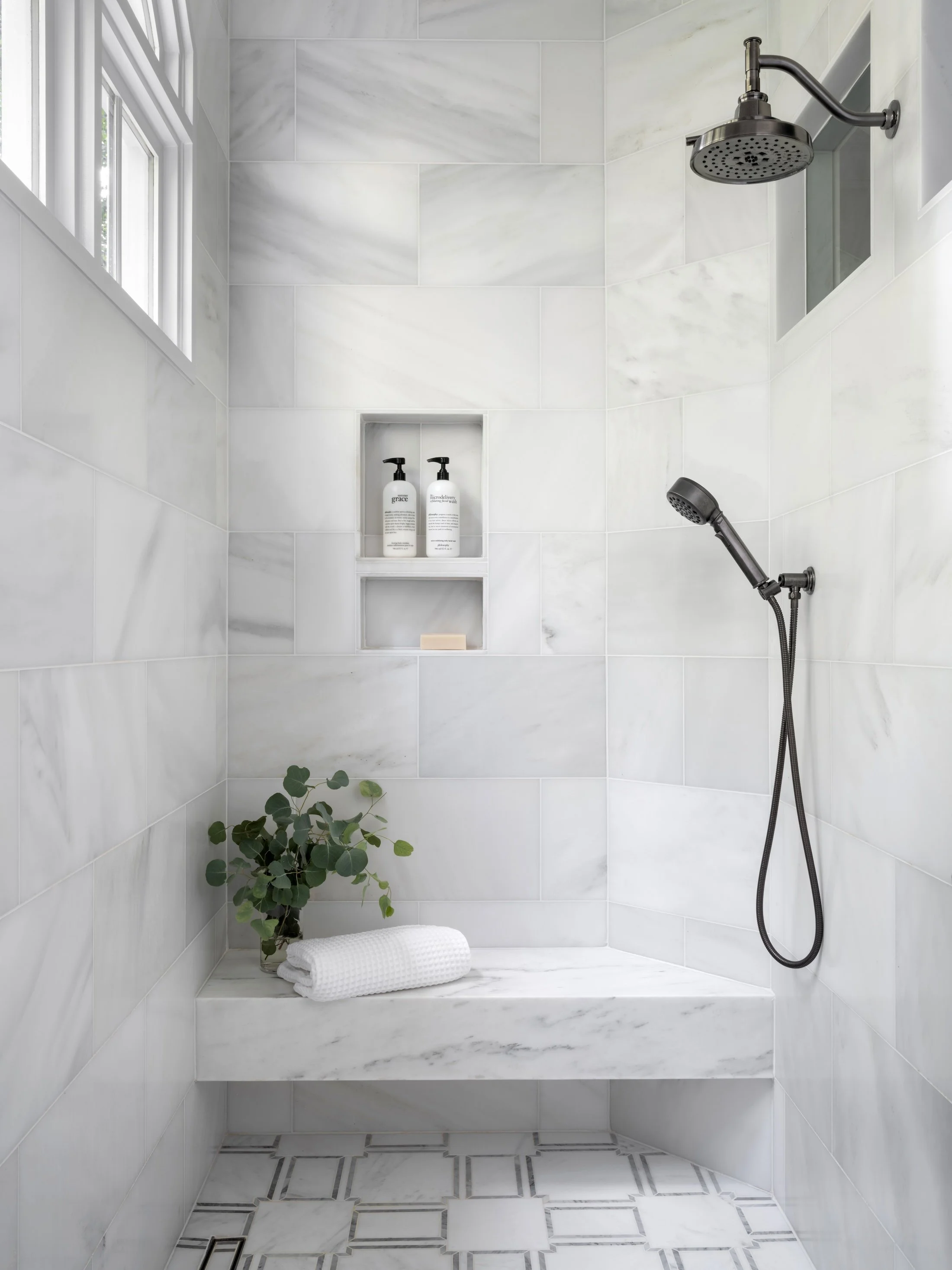 Marble walk-in shower with a built-in bench, recessed niche, handheld shower fixture, and soft natural light.