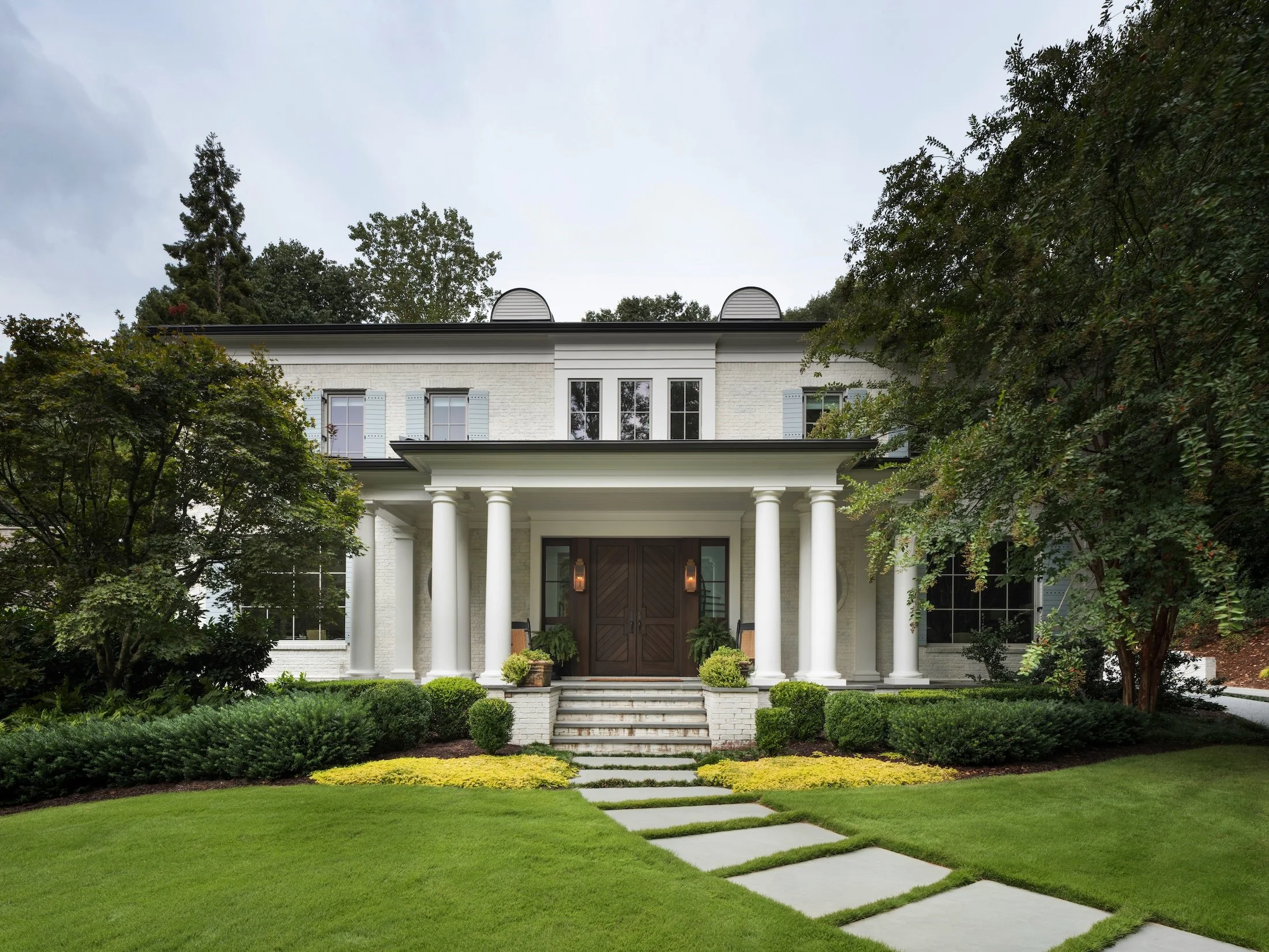 White brick two-story home with columned front porch, dark wood double doors, manicured landscaping, and a stepping-stone walkway.