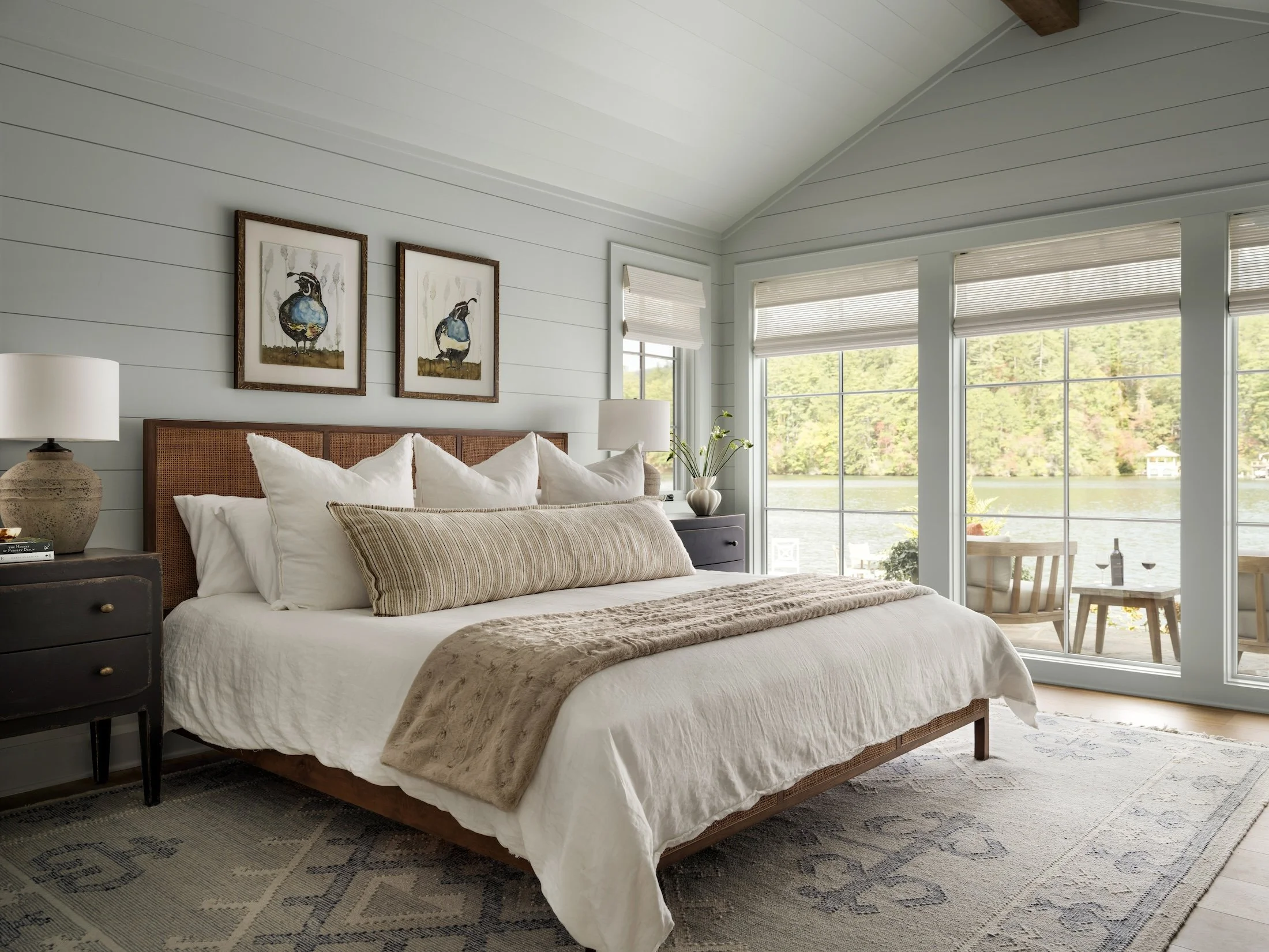 Serene bedroom with layered neutral bedding, woven headboard, dark nightstands, framed artwork, and soft natural light.
