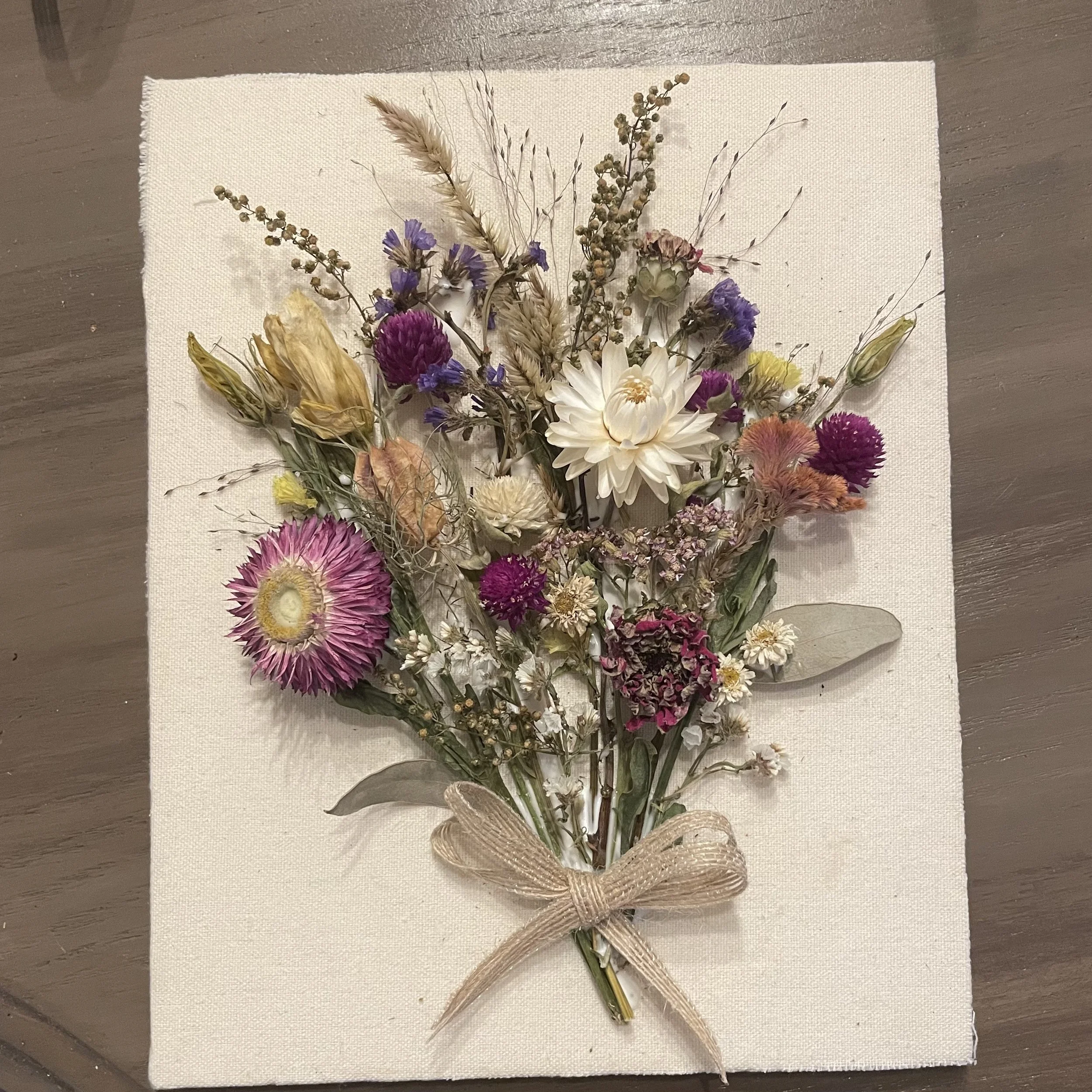 A bouquet of dried flowers with a beige ribbon tied around the stems, placed on a white textured cloth on a wooden surface.