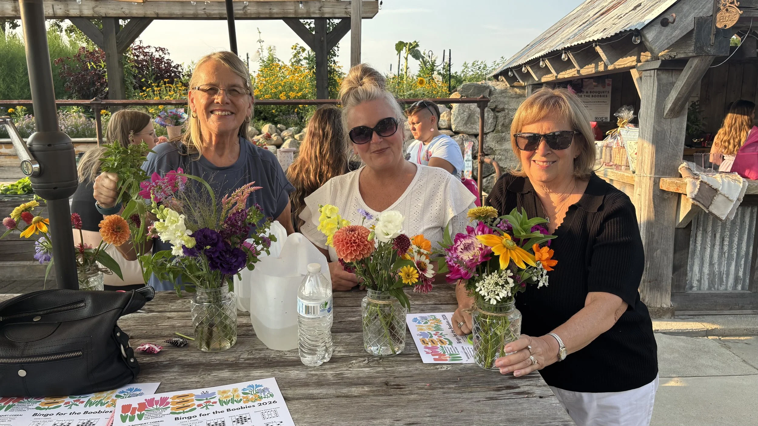 Three women sitting at a wooden table outdoors, smiling and holding colorful flower bouquets, with two water bottles and event flyers on the table, during a sunny day.