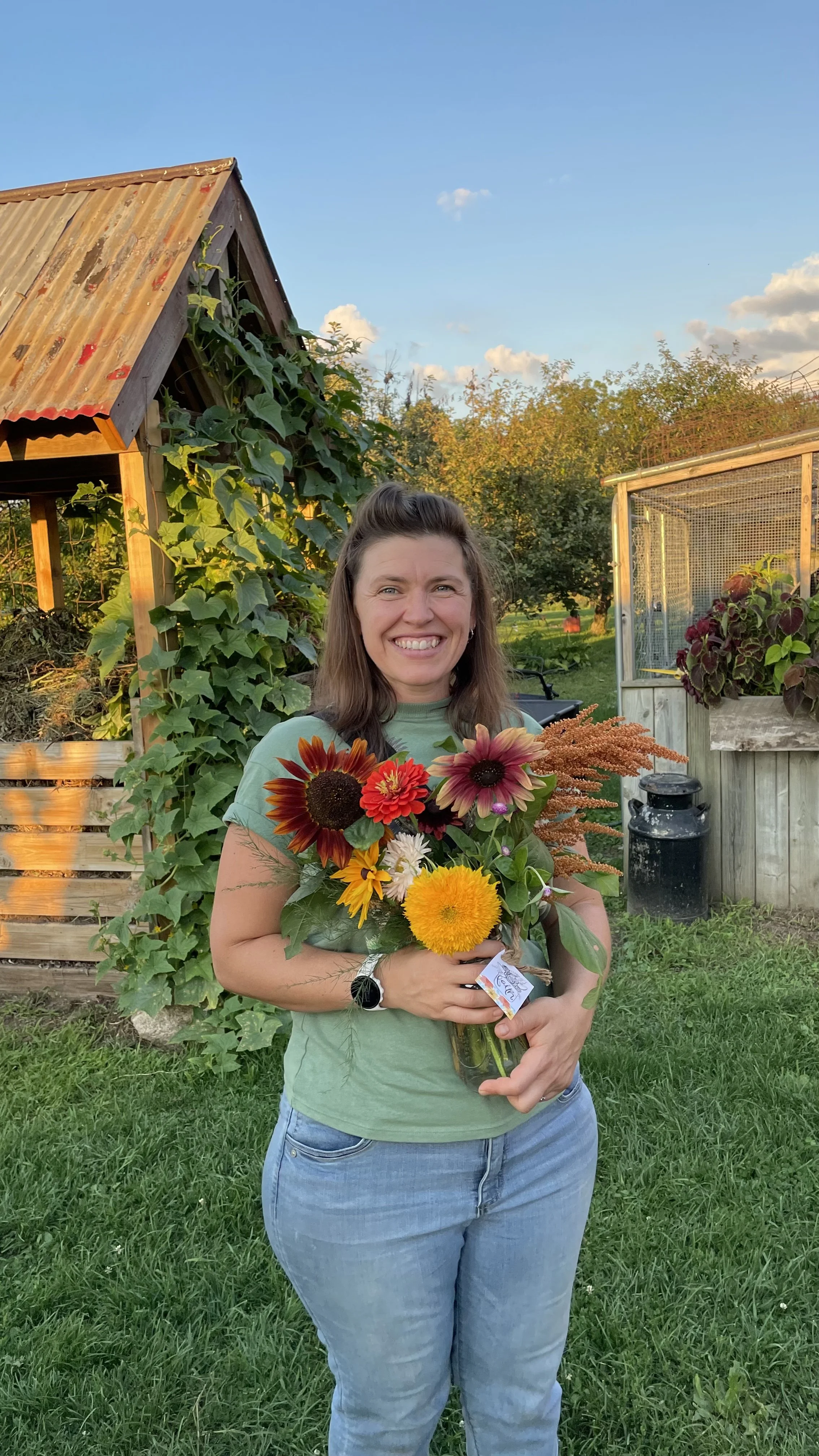 A woman smiling and holding a bouquet of colorful flowers, standing in a garden with greenery and a wooden structure in the background during sunset.