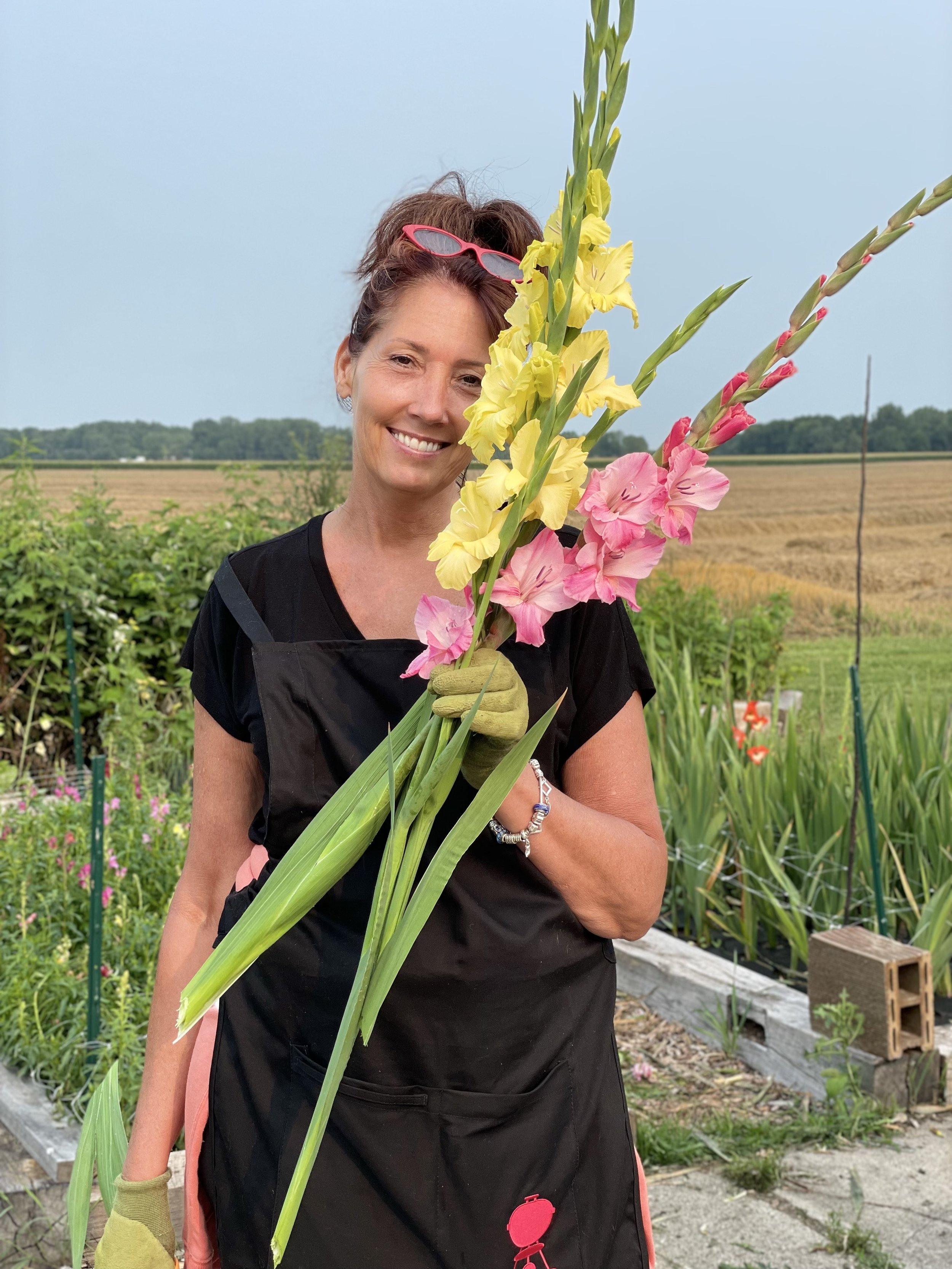 A woman smiling holds a large bouquet of colorful gladiolus flowers, wearing gardening gloves and a black apron, standing in a garden with plants and fields in the background.