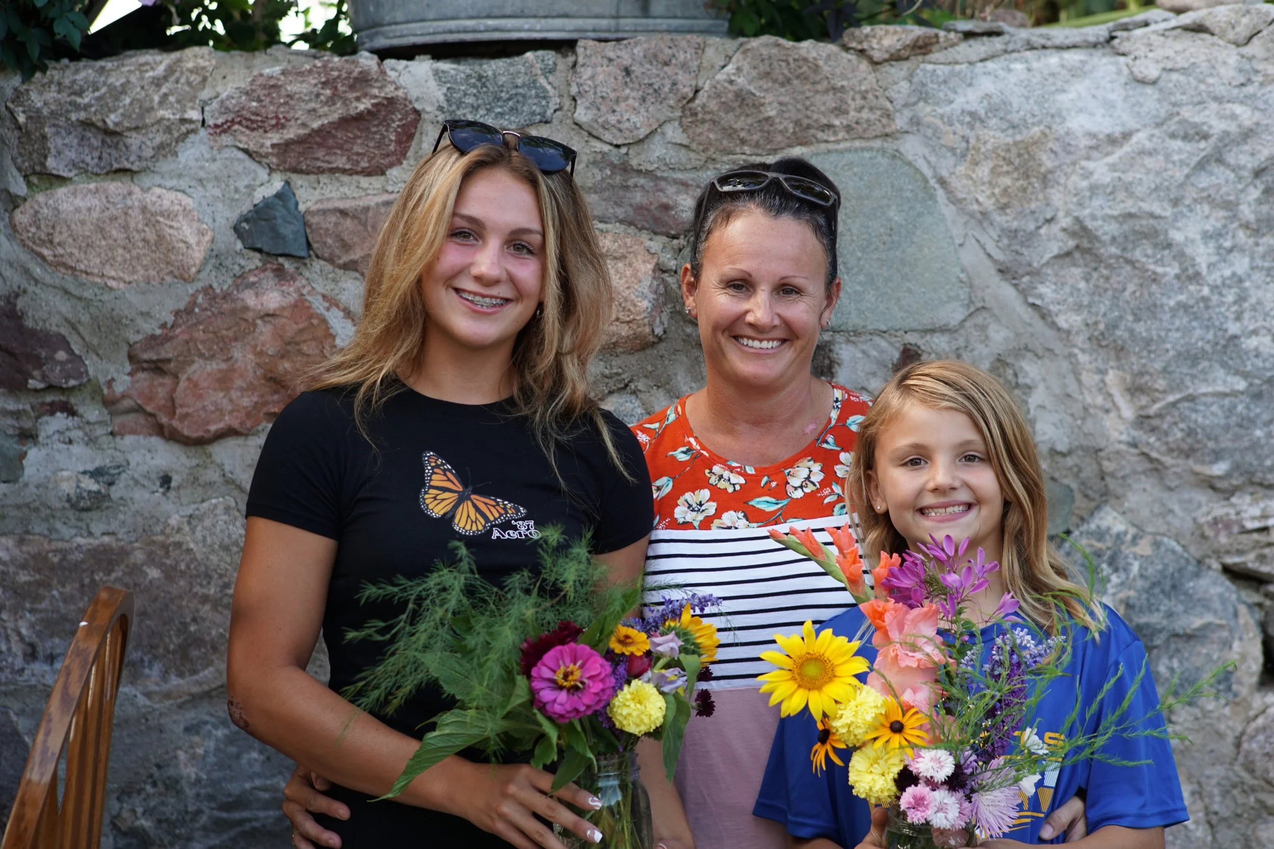A woman with sunglasses on her head, a girl with glasses, and a woman with her hair in a bun, all smiling while holding colorful flower bouquets, standing in front of a stone wall.