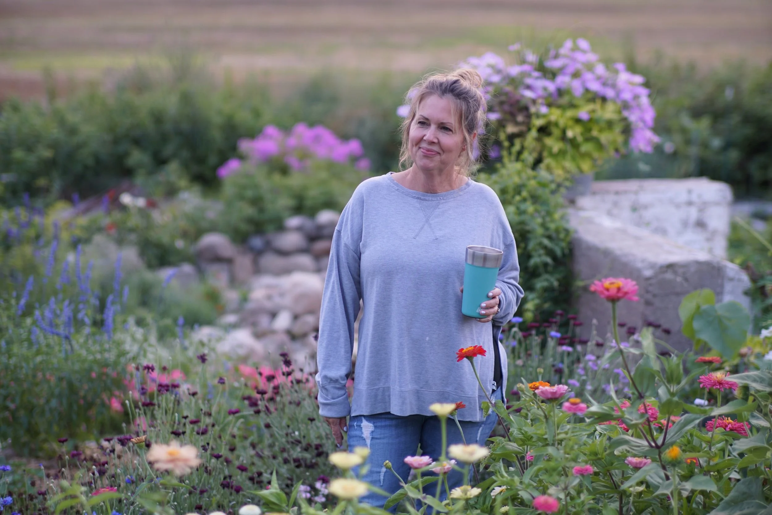 A woman with blonde hair in a messy bun, wearing a gray sweatshirt, smiles gently while holding a teal thermos, standing in a lush garden filled with colorful flowers and greenery.