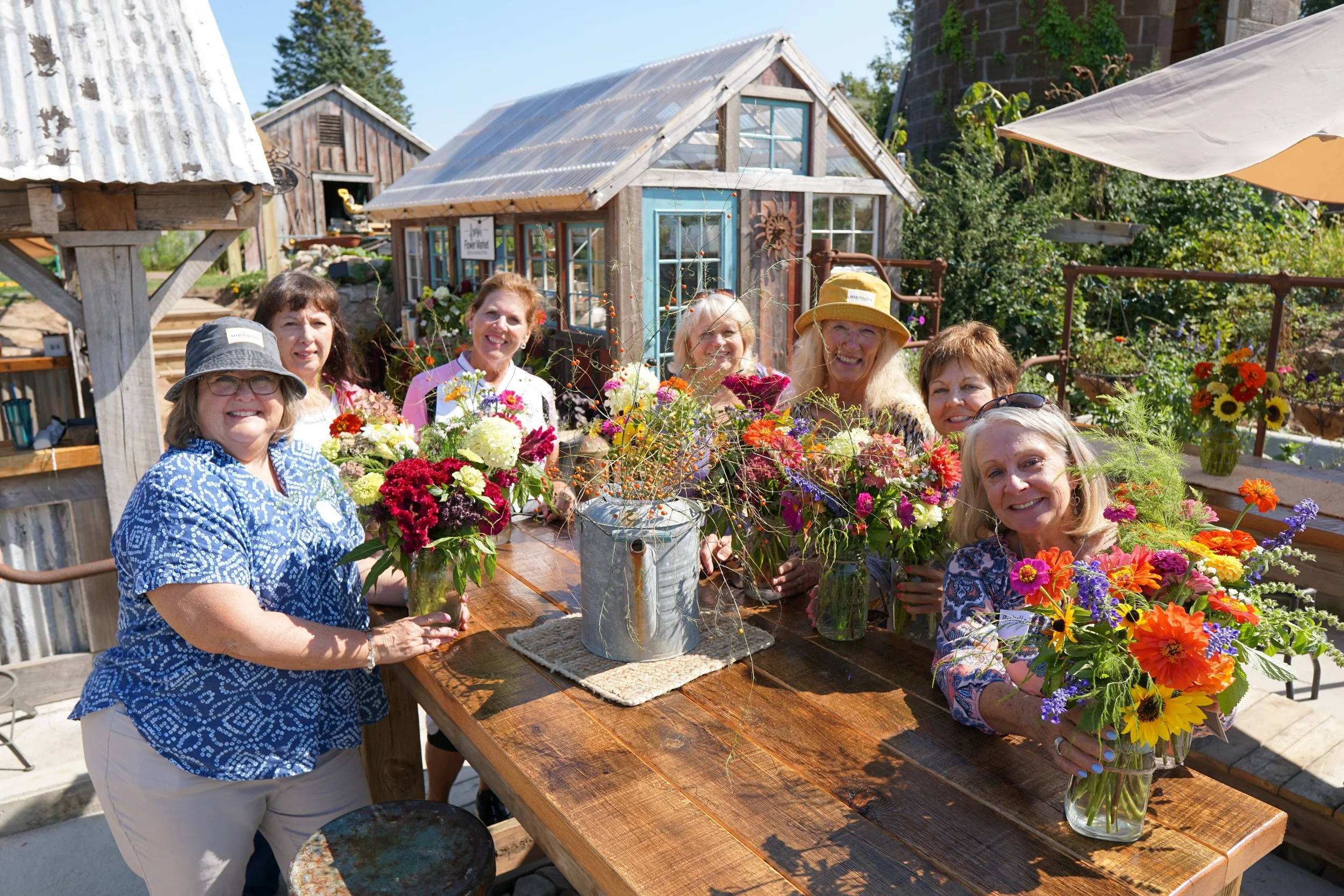 A group of women gathered around a large wooden table outdoors, holding colorful flowers in vases, with a rustic greenhouse and garden in the background.