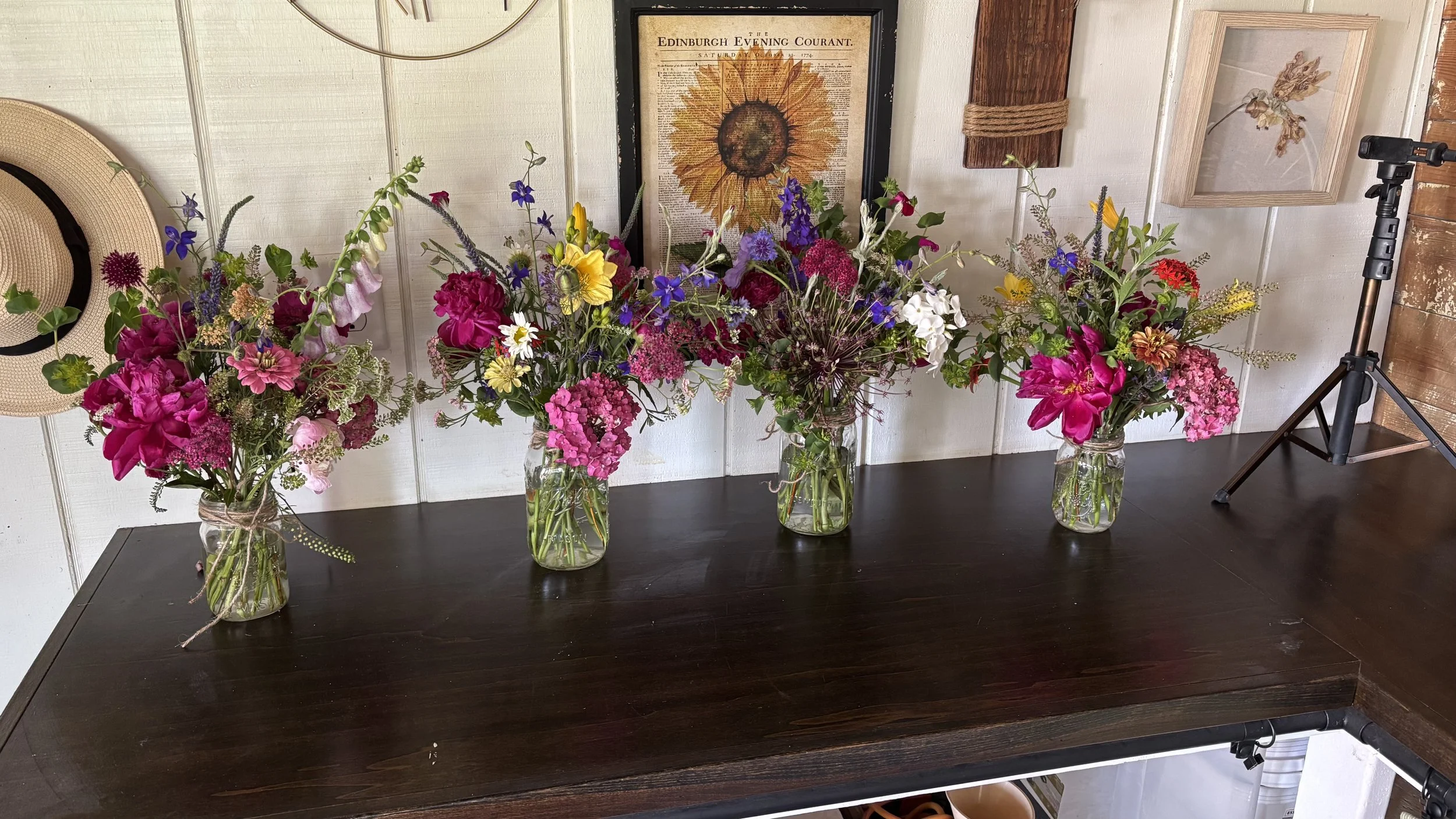 Four glass jars filled with colorful mixed flowers, placed on a dark wooden table against a white paneled wall decorated with framed artwork and a hat.