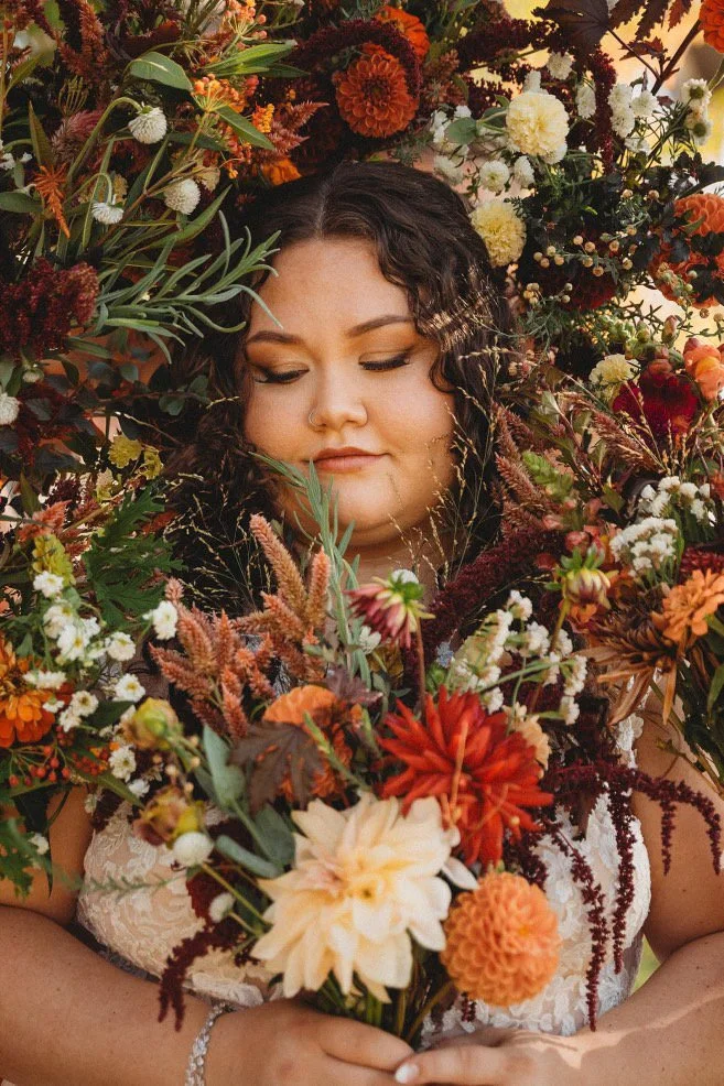 A woman with dark, curly hair and closed eyes holding a large bouquet of colorful flowers amidst a wall of various flowers and foliage.