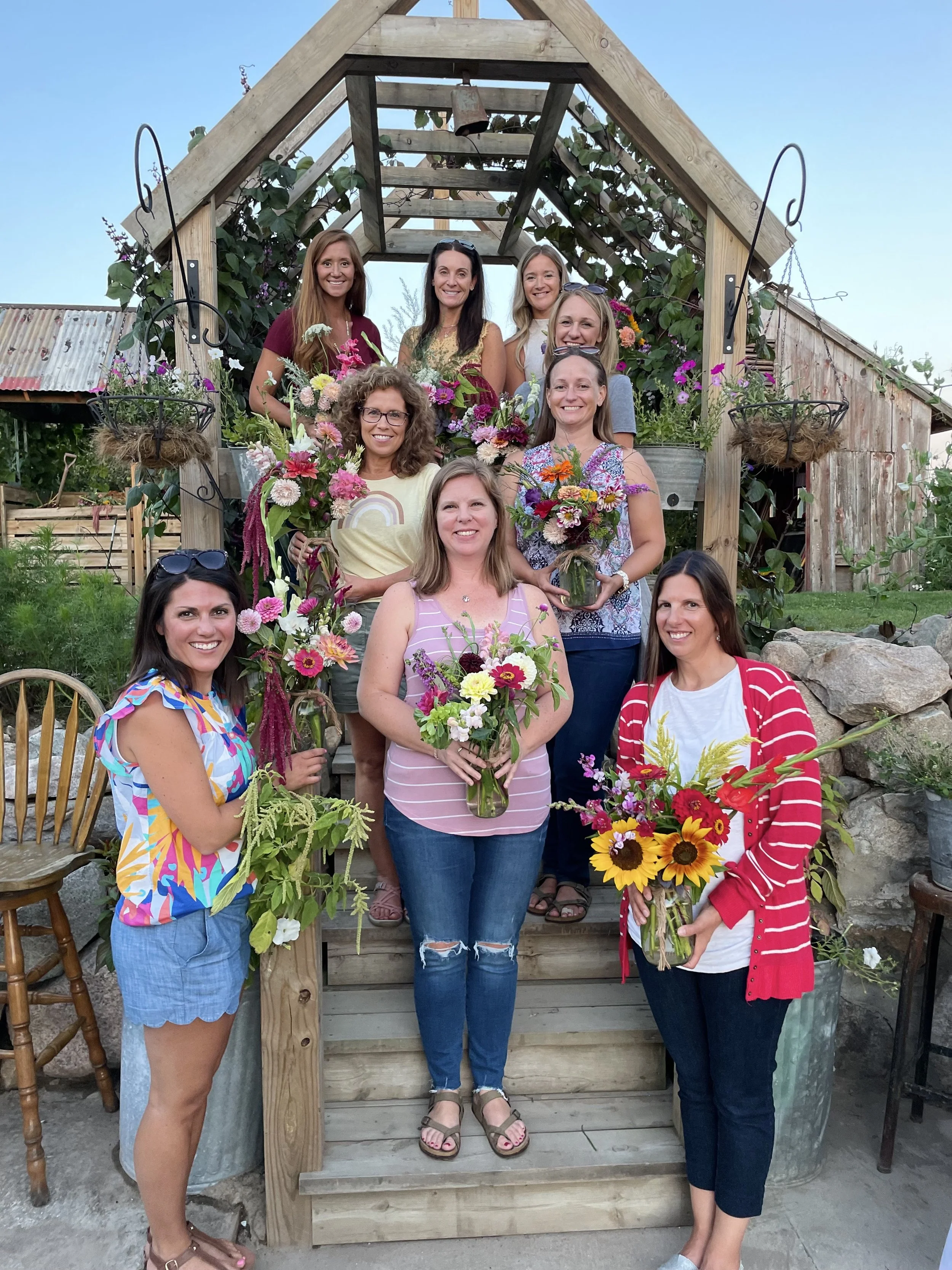 A group of nine women standing on a wooden staircase, holding colorful bouquets of flowers, in an outdoor garden setting with a wooden shed and plants in the background.