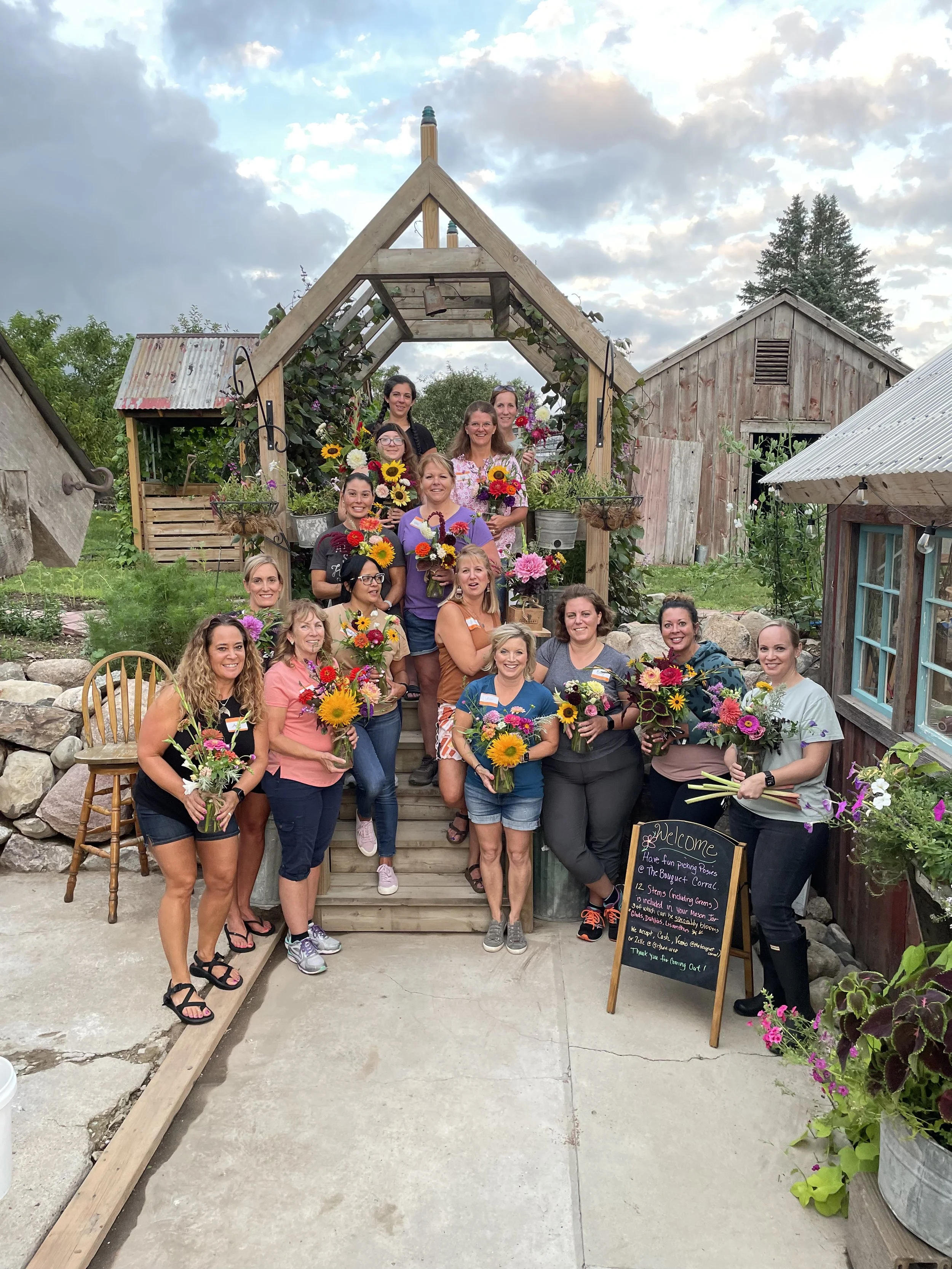 Group of women holding flower bouquets standing on the steps of a wooden garden or nursery with rustic barns and plants around, overcast sky in the background.
